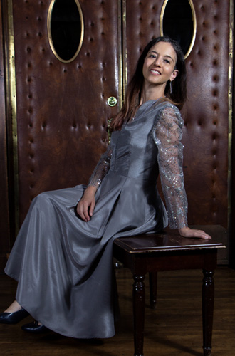 Young woman in a long, elegant gray dress sitting on a small wooden table, smiling, with a wooden door background.