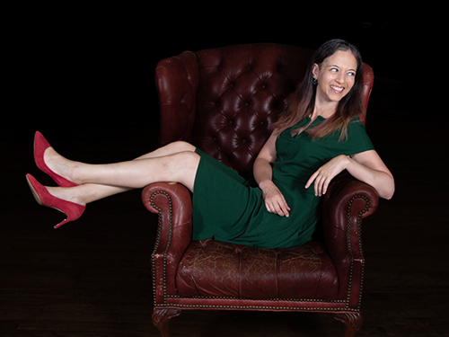 Woman in green dress and red heels sitting on a leather armchair, smiling, against a black background.