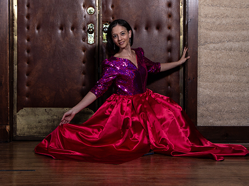 Young girl in a purple and red gown sitting on the floor in front of a large door, smiling.