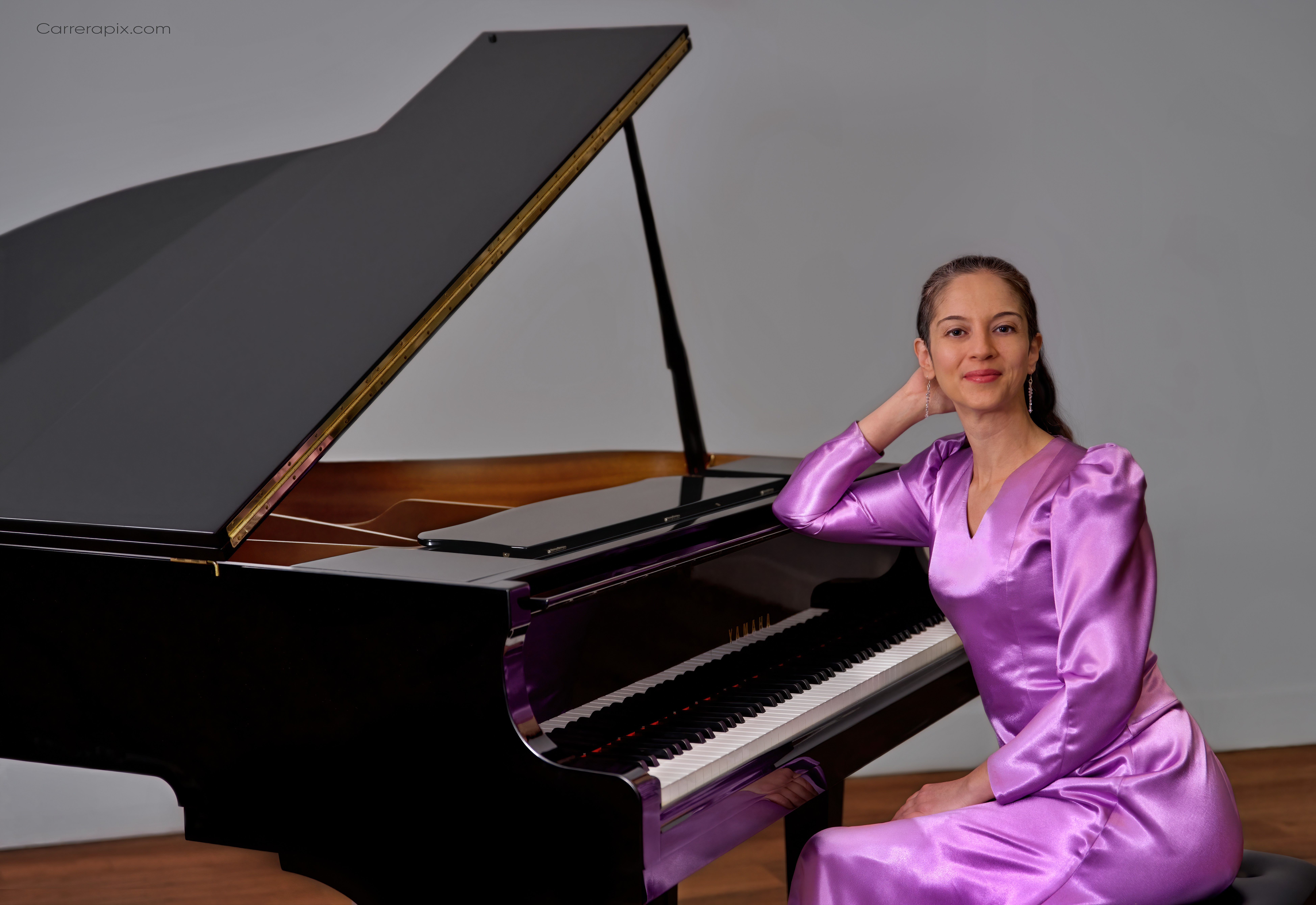 Woman in purple dress sitting beside a grand piano with open lid, smiling, gray background.