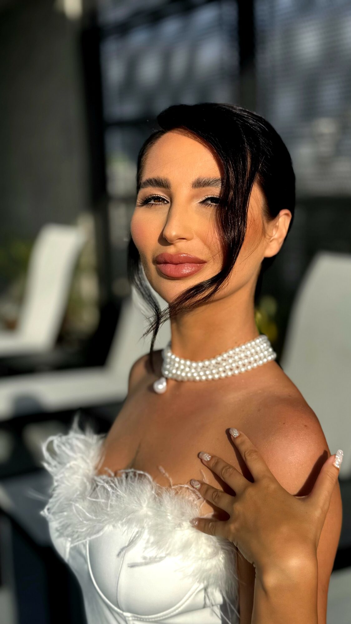Woman with dark hair, wearing pearl necklace and white dress with feather details, outdoors with chairs in background.