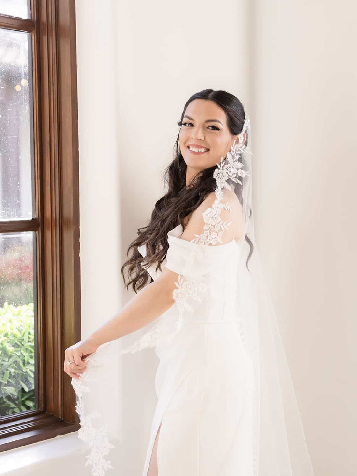 Smiling woman in a white dress with lace details, standing indoors near a window, holding her dress, with long dark hair.