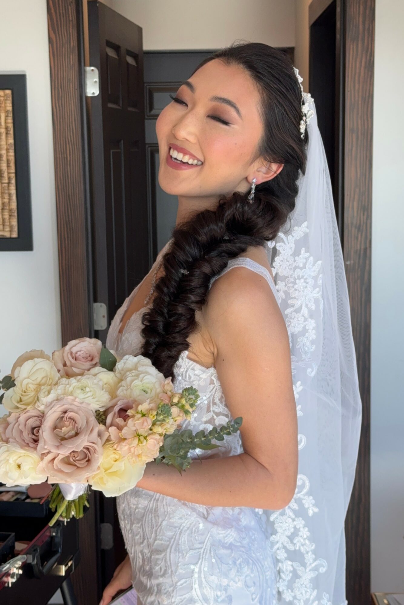 Smiling bride with long braided hair, holding a bouquet of roses, wearing a lace wedding dress and veil, standing indoors.