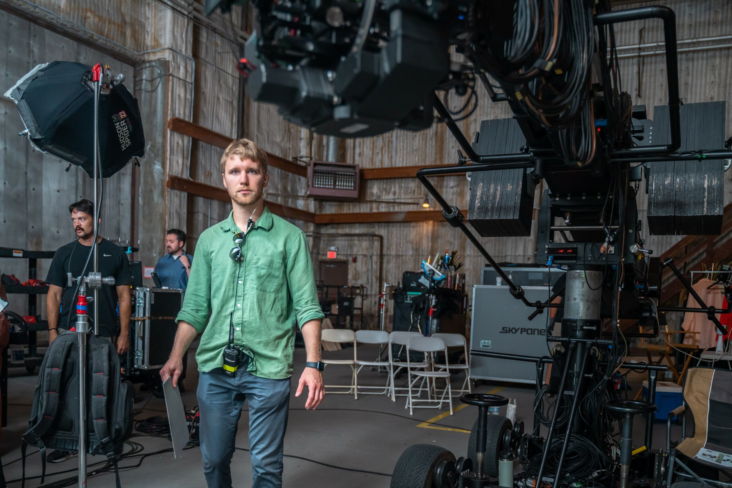 People working on a film set with camera equipment and lighting, a man in a green shirt in foreground.