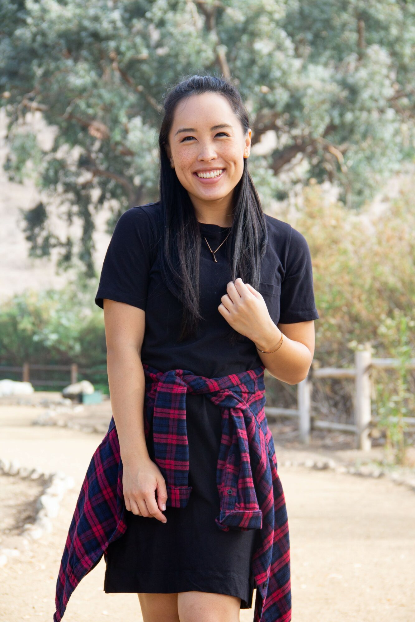Young woman smiling outdoors, wearing a black dress and a red plaid shirt tied around her waist, with trees in background.