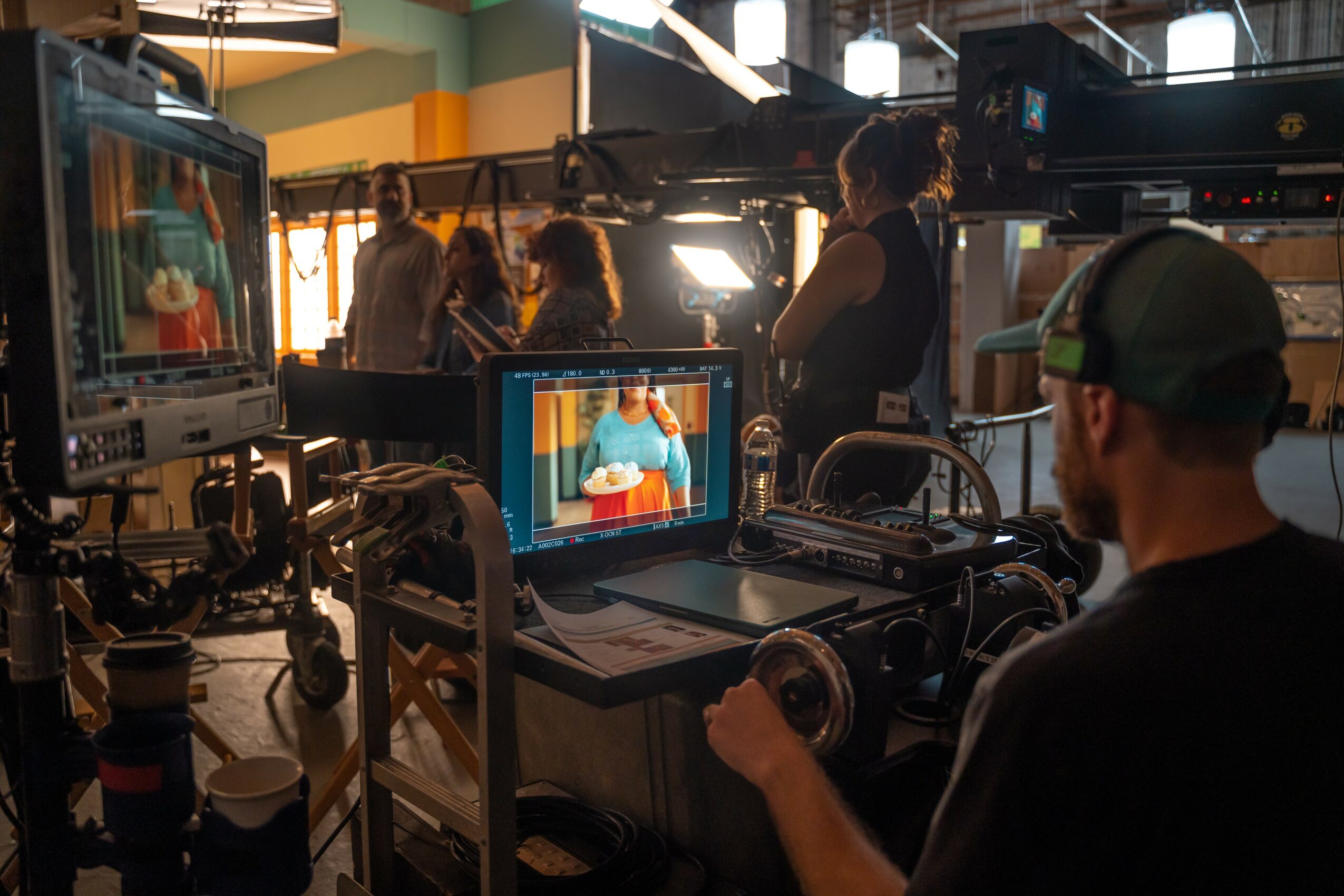 A person operating a camera in a studio with crew and equipment, filming a person on a monitor, with lighting and set elements.