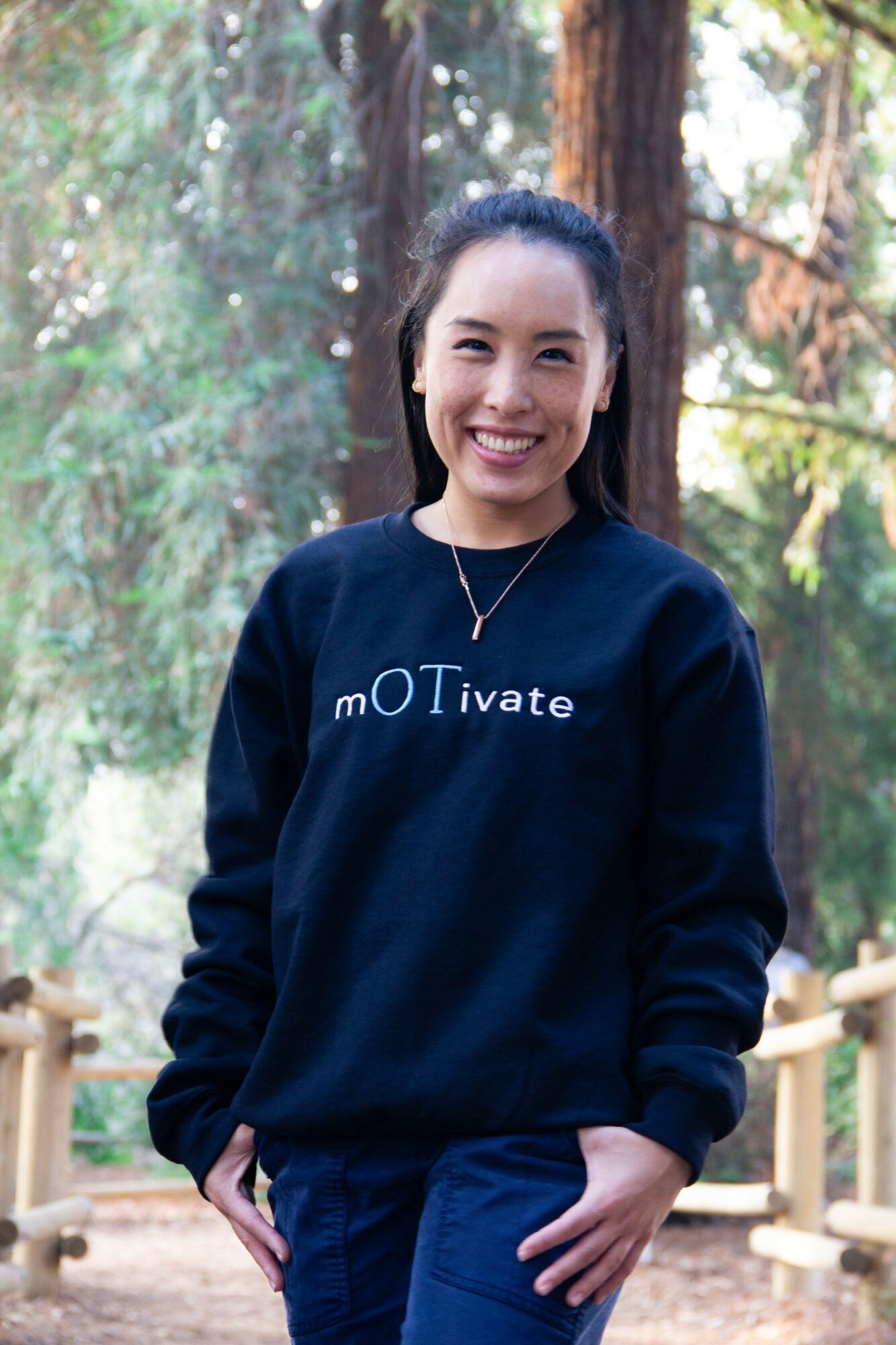 Young woman smiling outdoors in a forest, wearing a black sweatshirt with text, standing near a wooden fence.