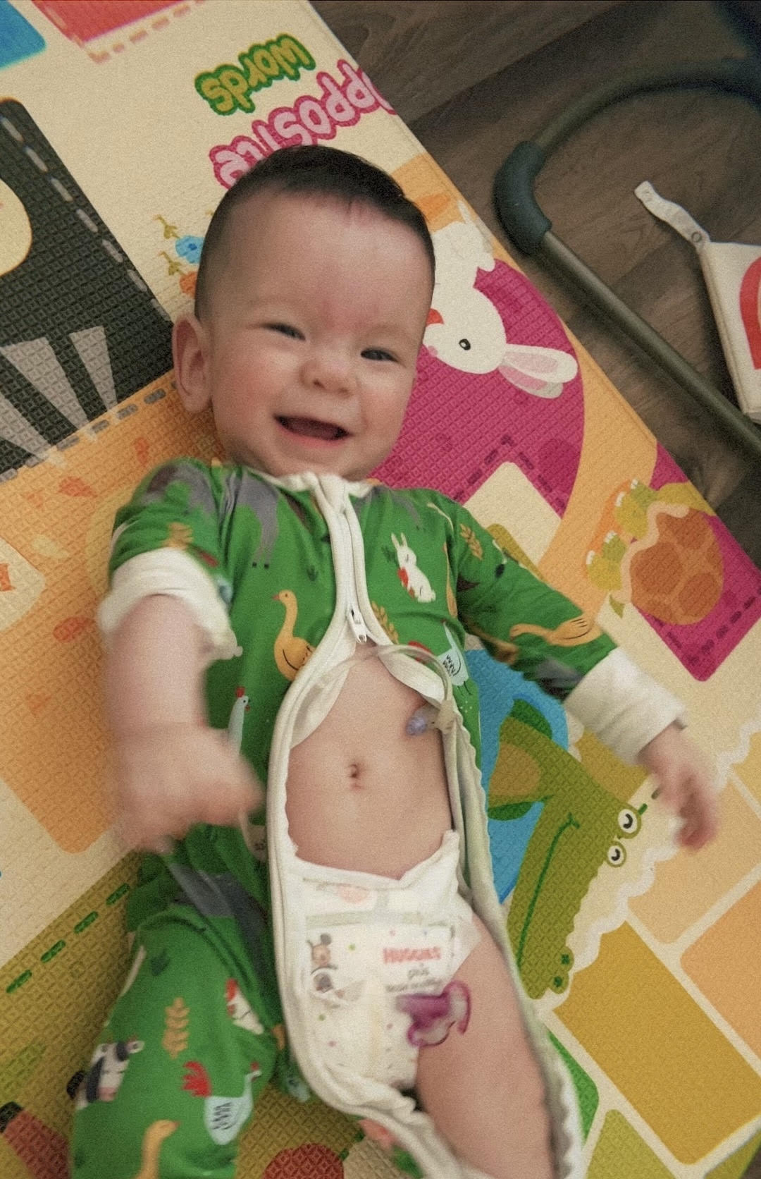 Smiling baby lying on colorful play mat, wearing green outfit, with diaper visible, in a cheerful setting.
