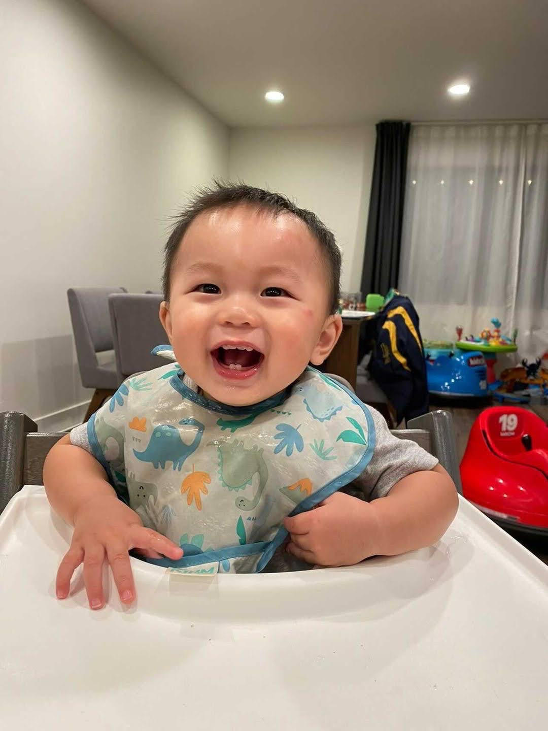 Smiling baby sitting in high chair with a colorful background, wearing a patterned shirt, indoors at night.