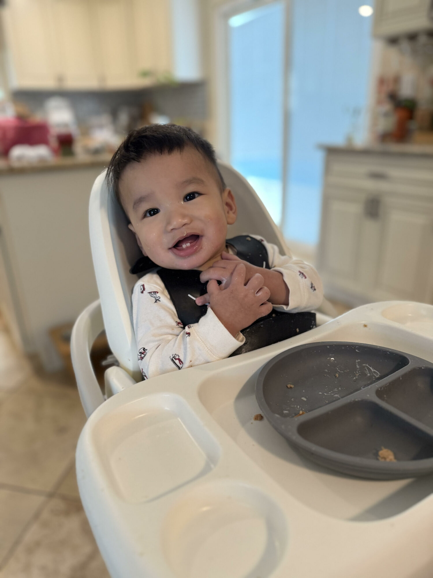 Smiling baby sitting in high chair in a kitchen, holding hands together, with a blue door in the background.