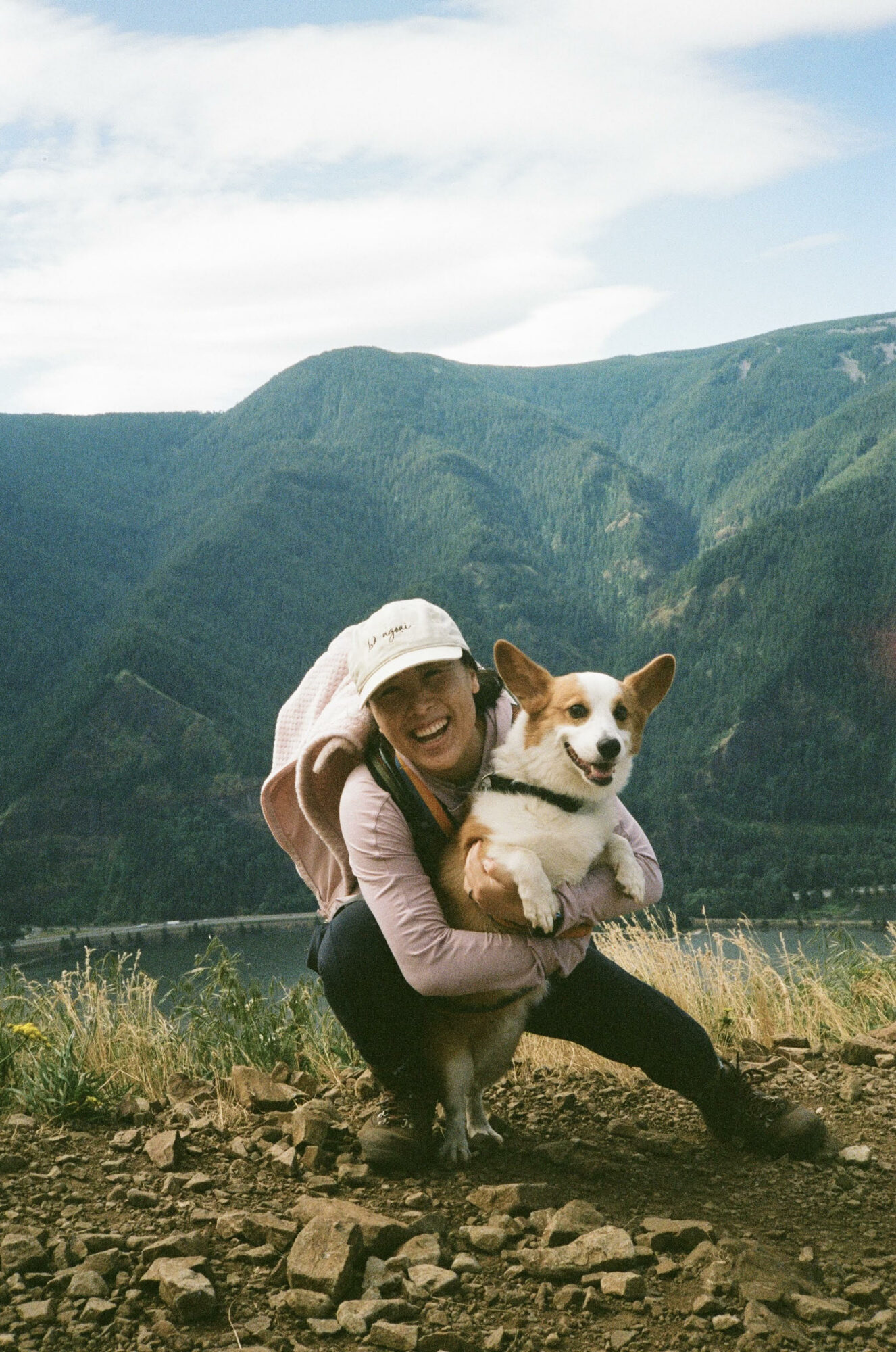 Person crouching outdoors with a dog against a mountainous landscape, blue sky, and grassy foreground.