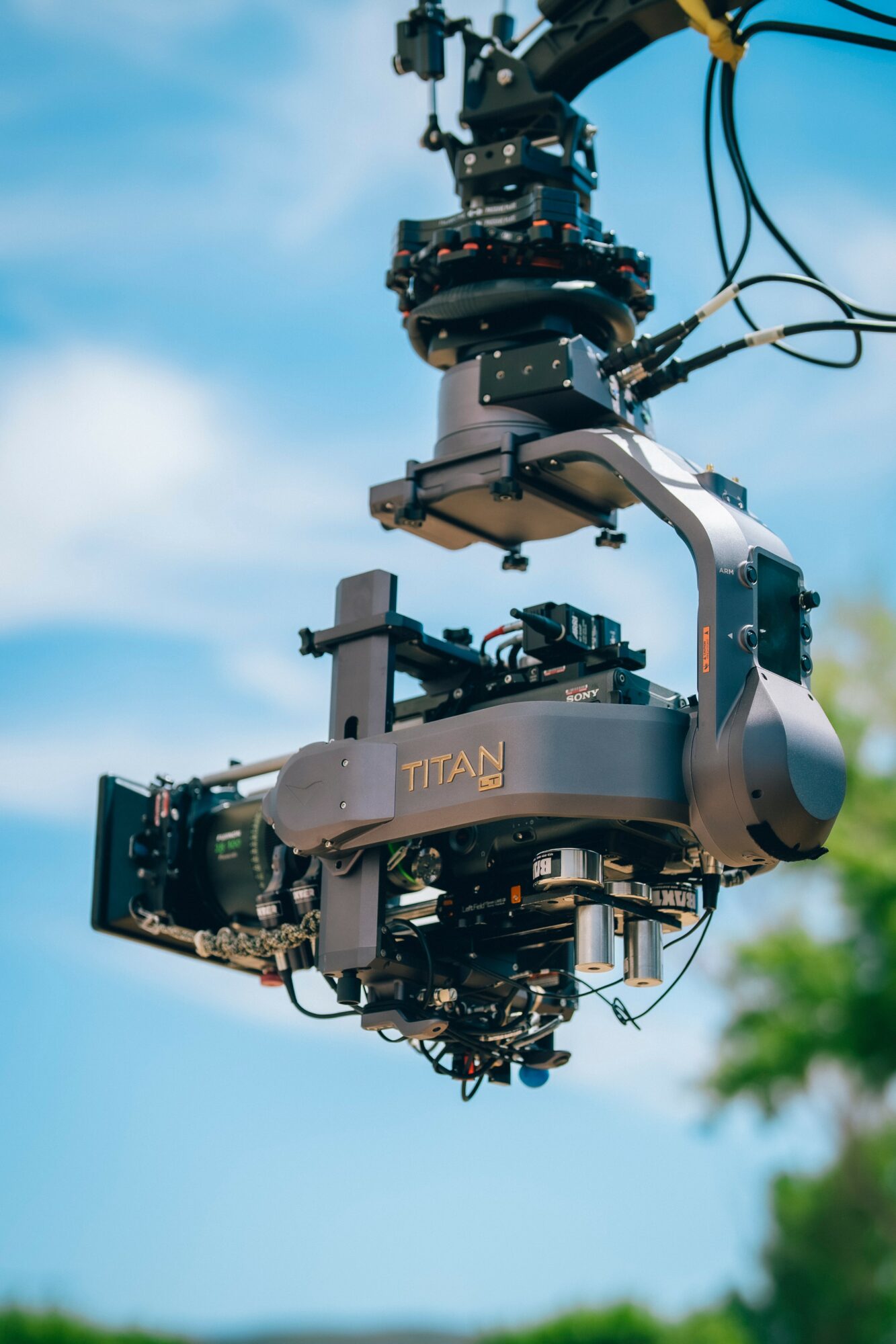 A drone with a camera mounted on a gimbal, labeled 'TITAN,' flying outdoors with a blue sky and green trees in background.