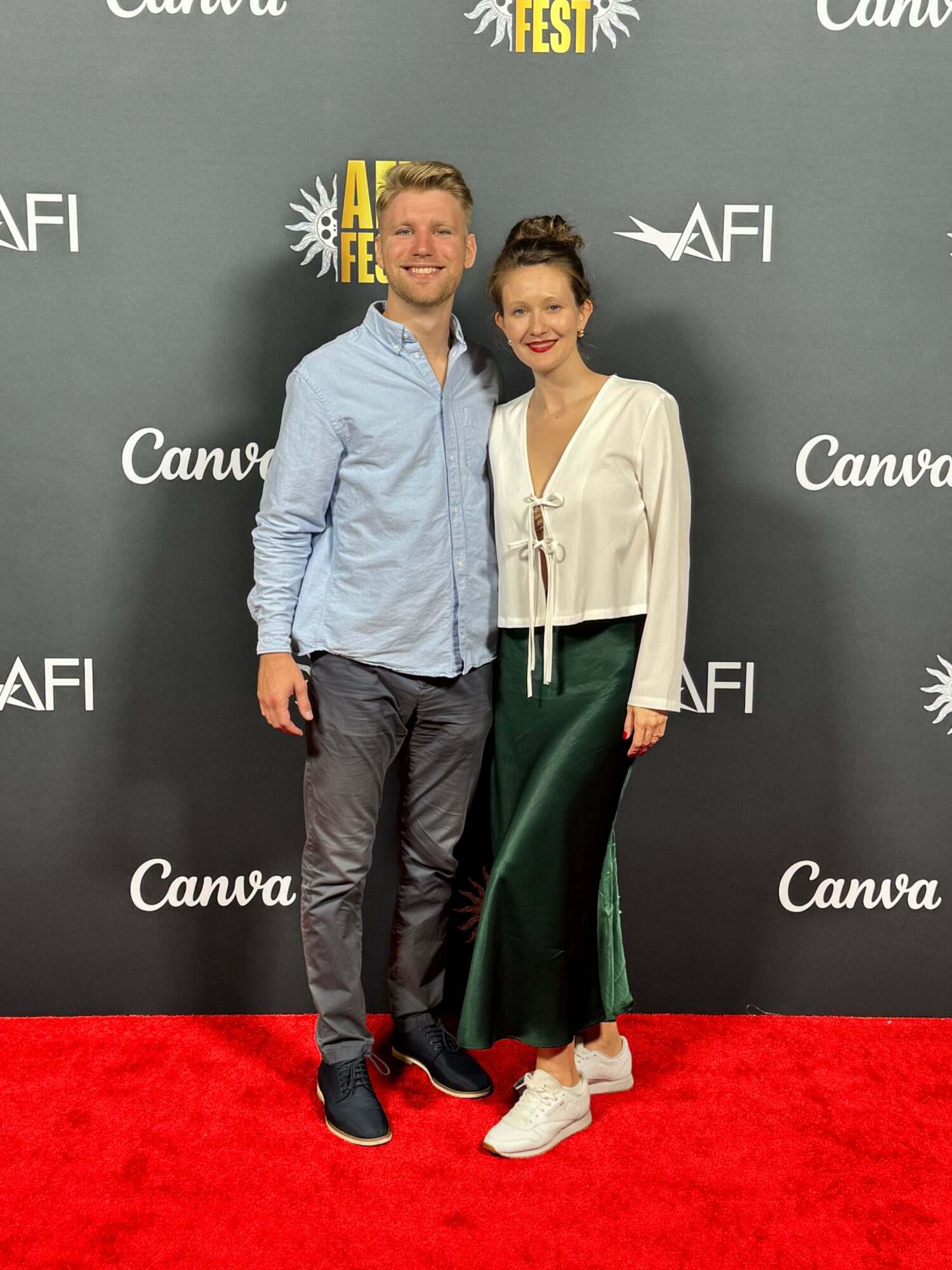 Two people standing on a red carpet in front of a black backdrop with white text, posing for a photo.