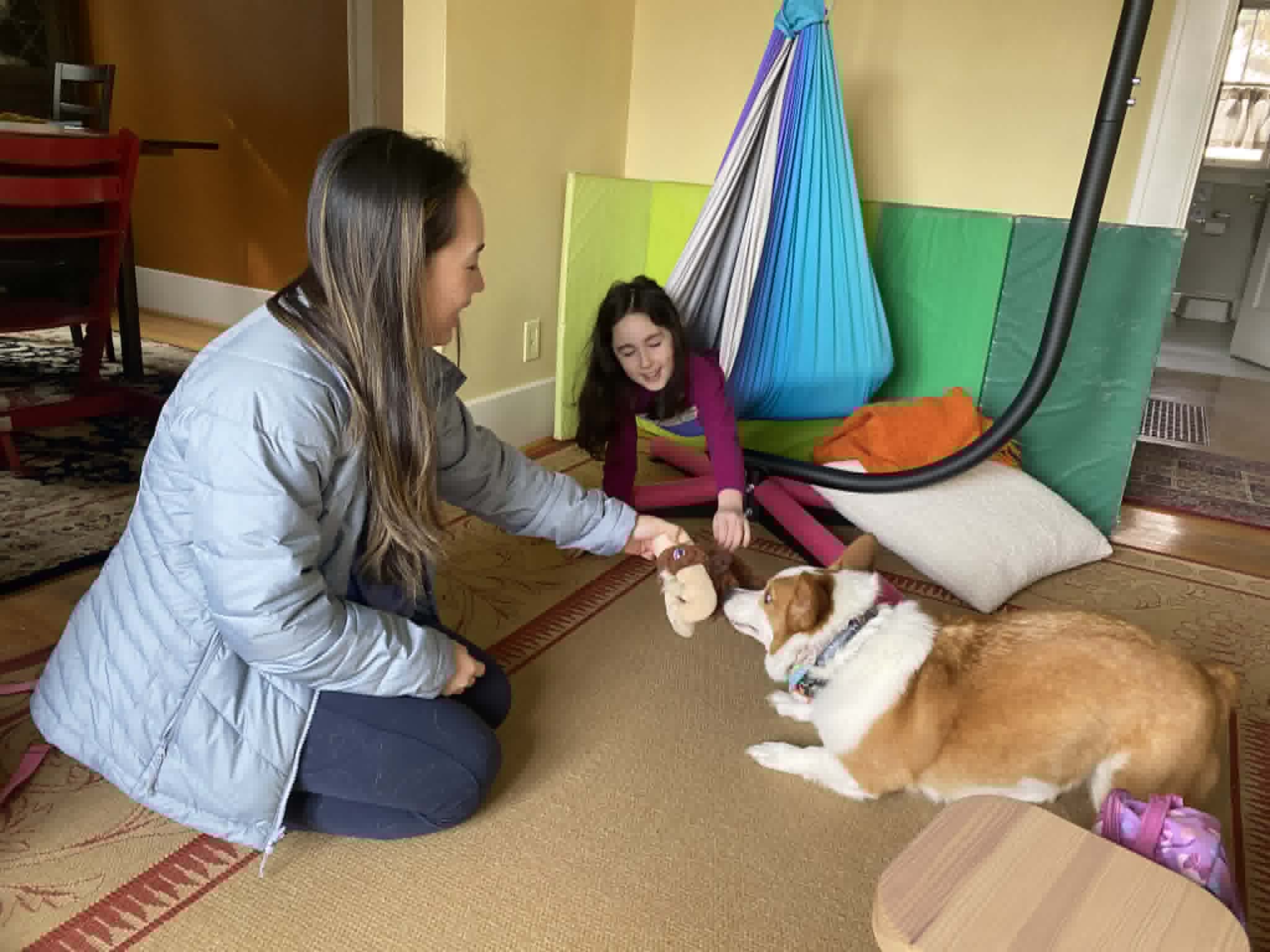 Woman and girl playing with a dog indoors, with colorful hanging fabric and cushions in the background.