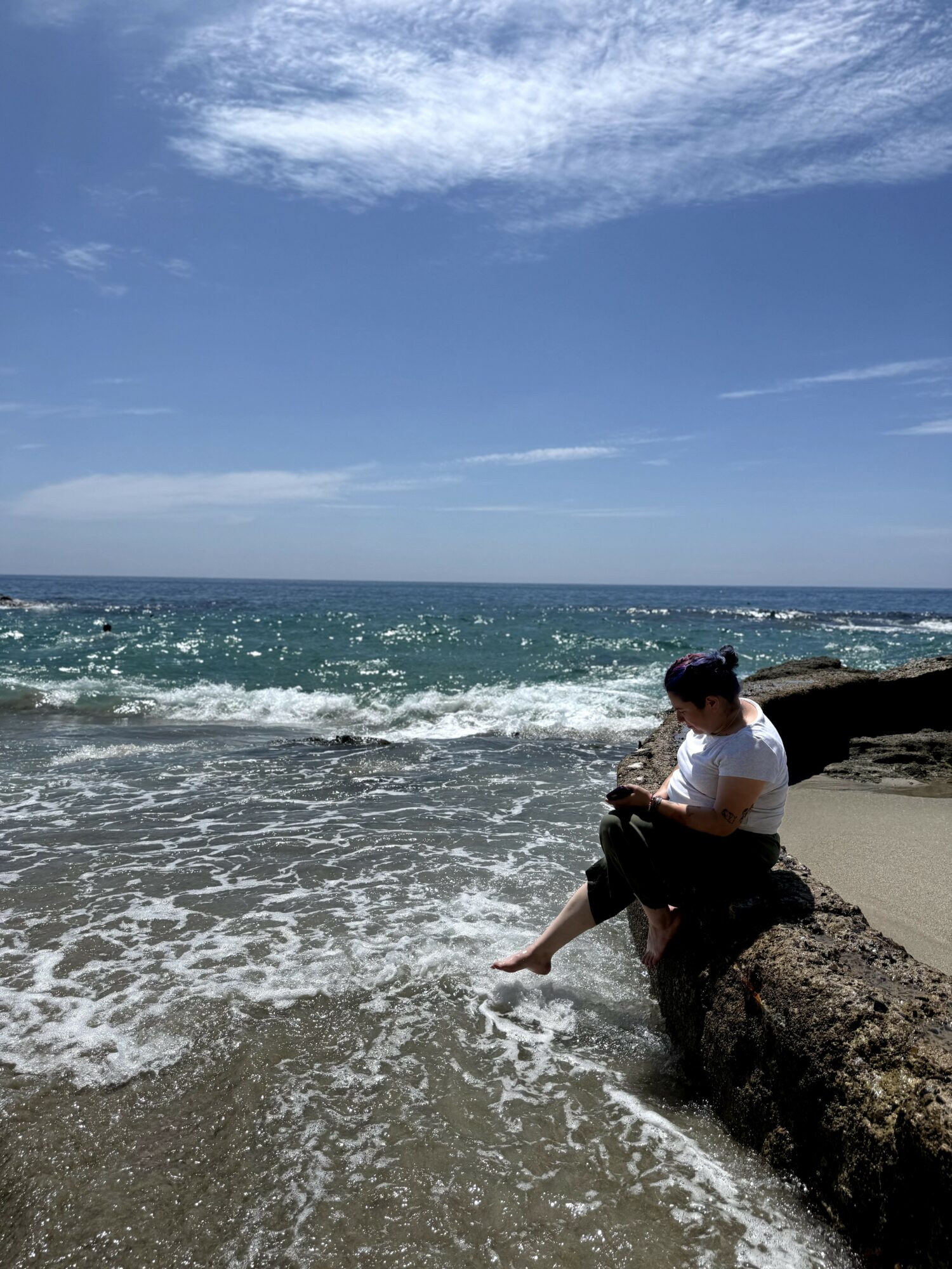 Person sitting on a log at the beach, with ocean waves and blue sky in the background.