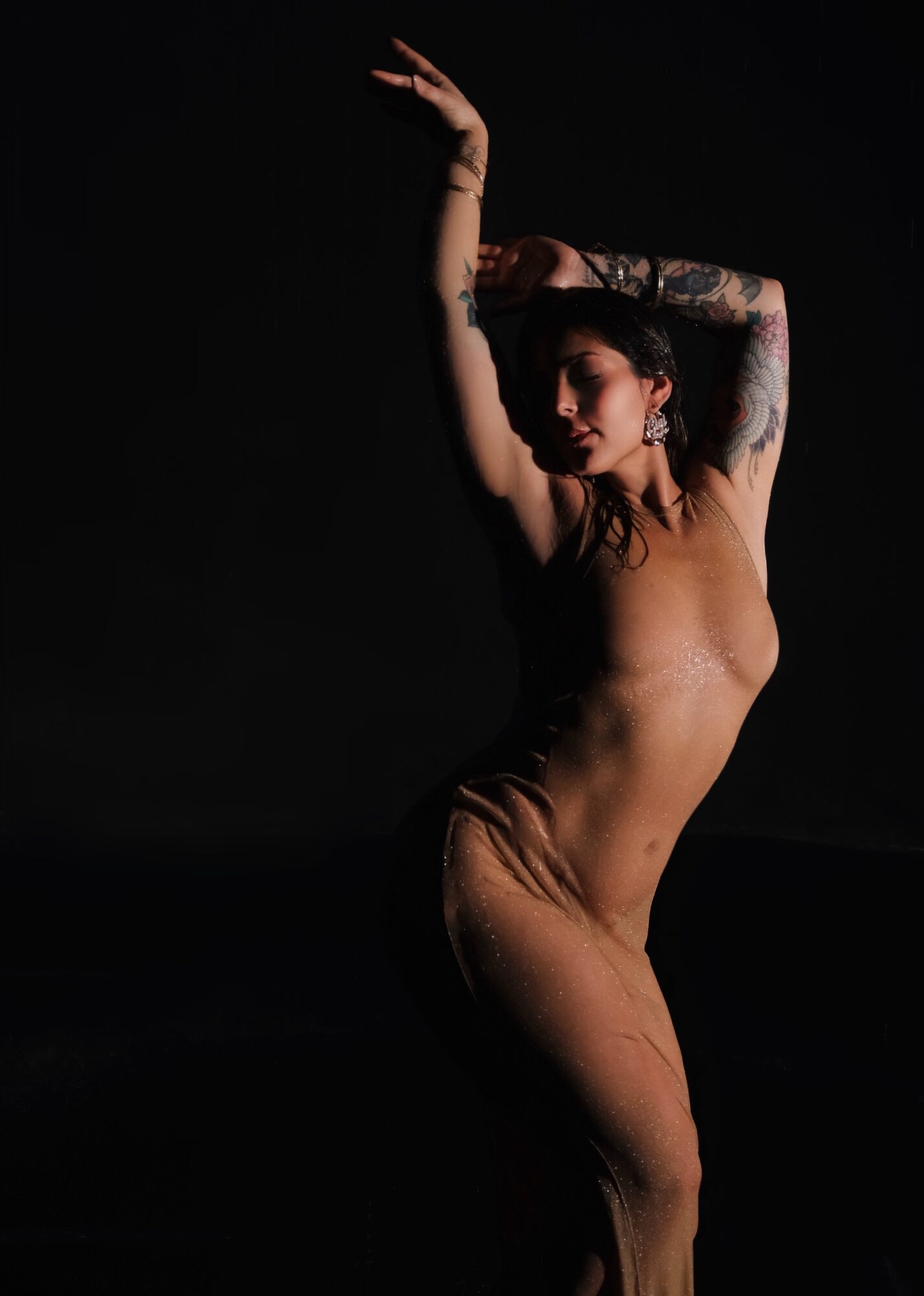 A woman with tattoos poses with one arm raised above her head against a dark background.