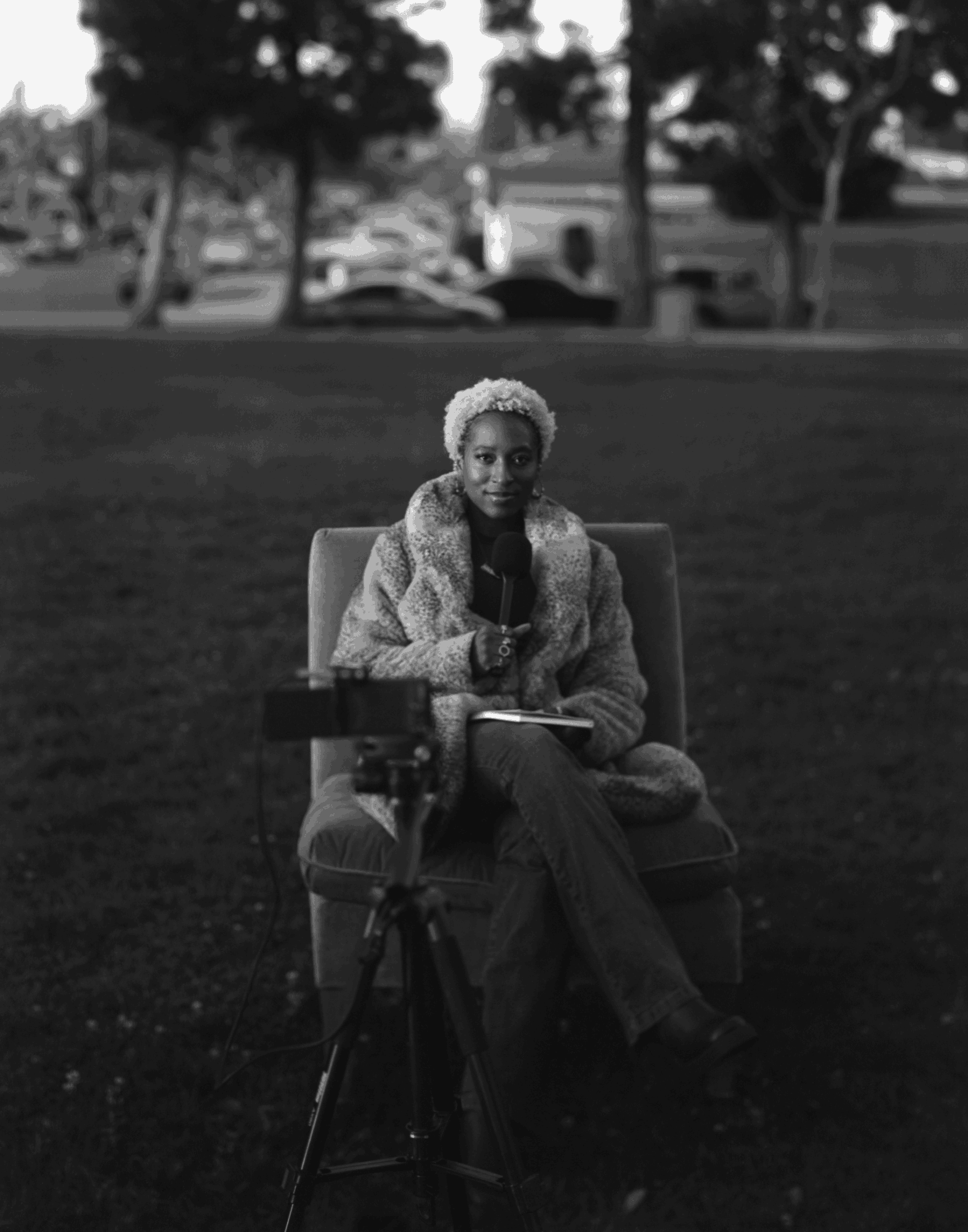 Woman with short curly hair sitting on a chair outdoors, holding a microphone, with trees and cars in the background.
