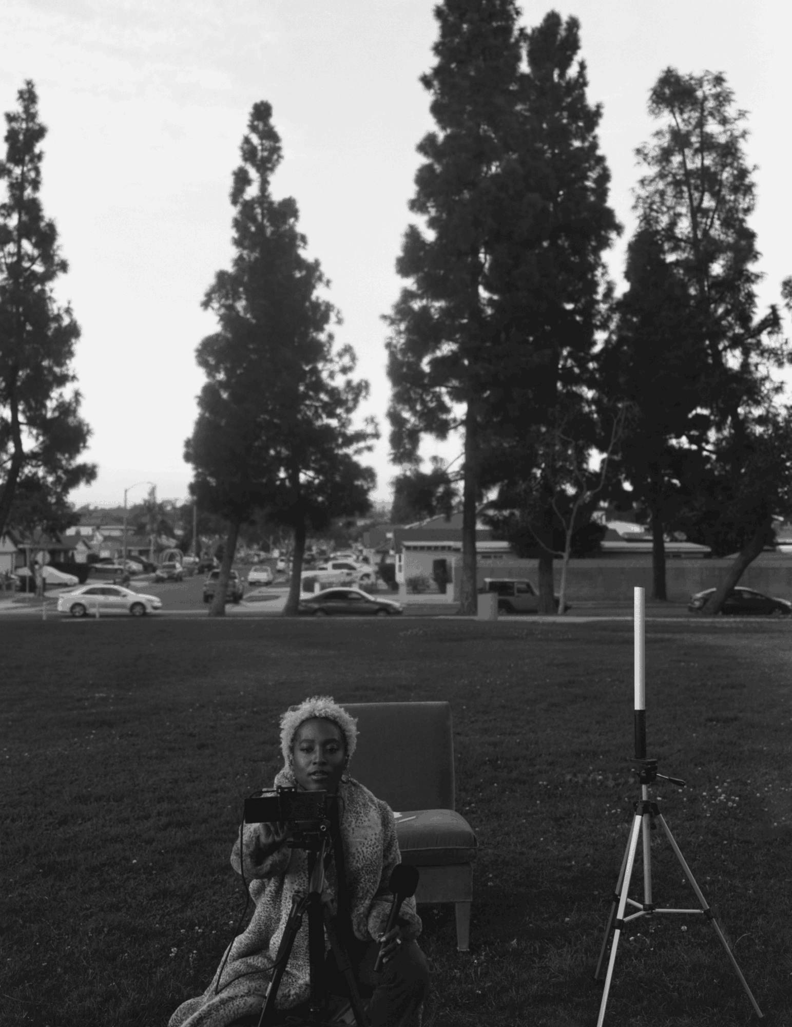 Person sitting on a chair outdoors with trees and parked cars in the background, holding a device, in black and white.