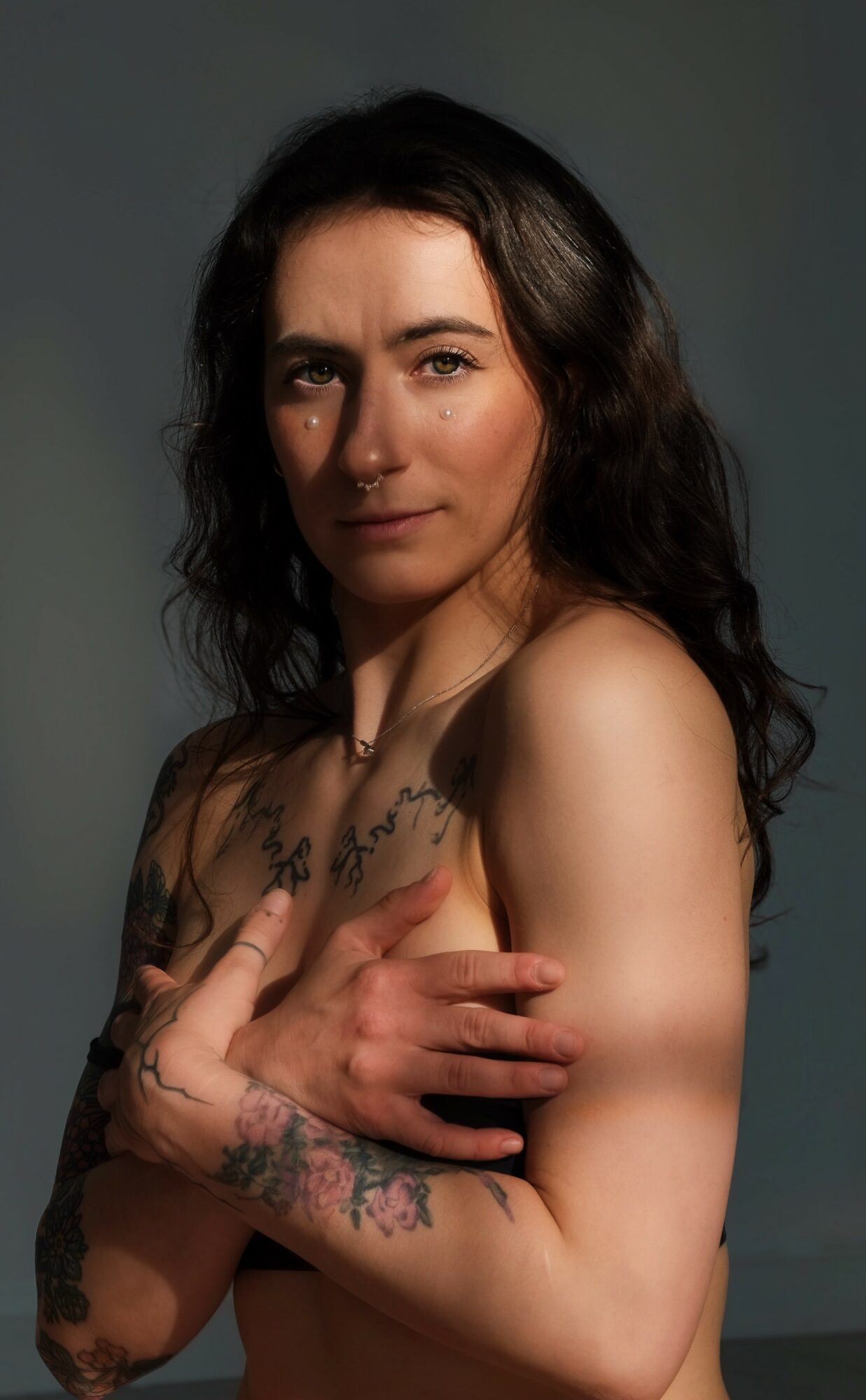 Woman with long dark hair, tattoos on arms and chest, looking at camera with a neutral expression.