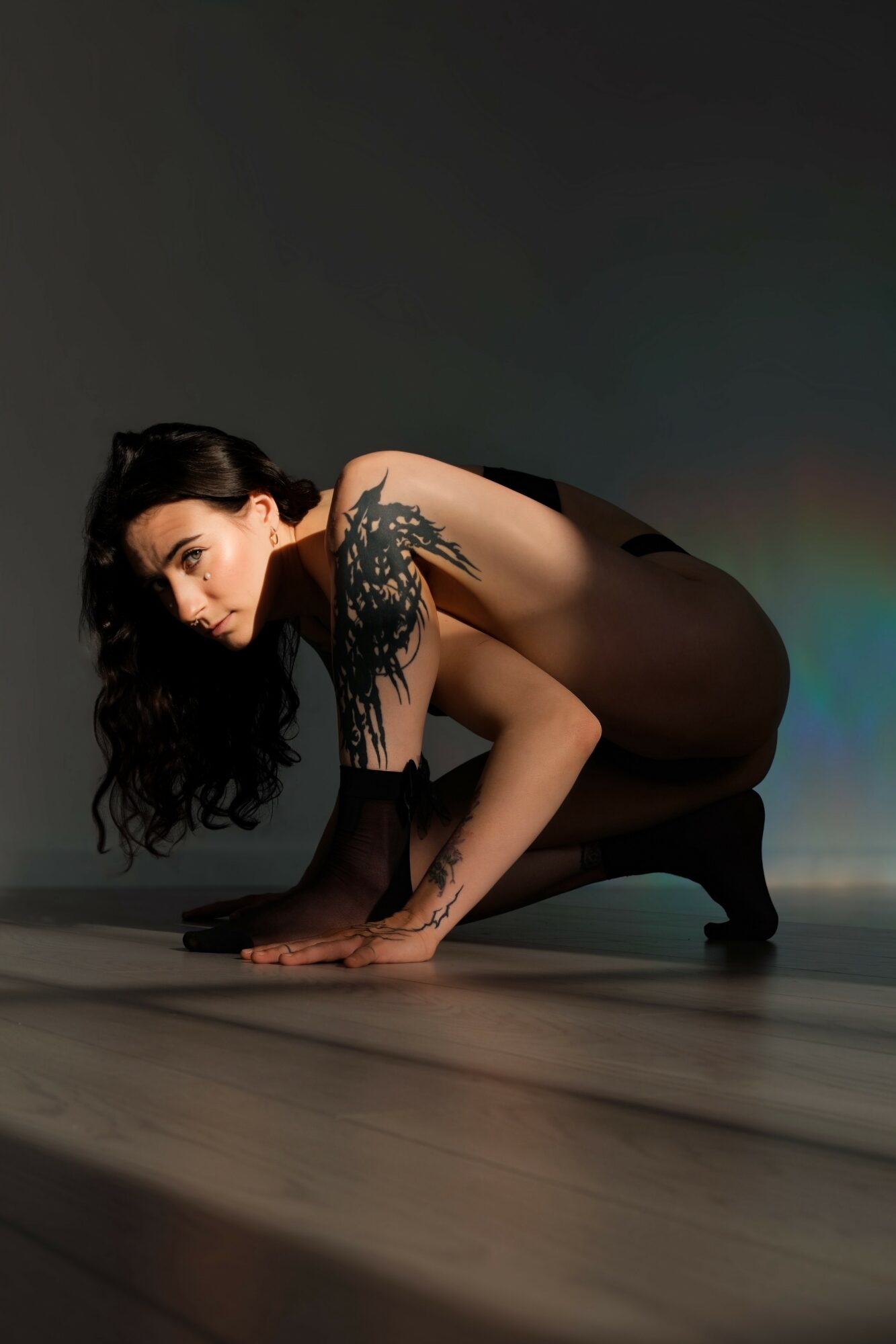 Woman crouching on floor with dark background, wearing black dress and showing a tattoo on her shoulder.