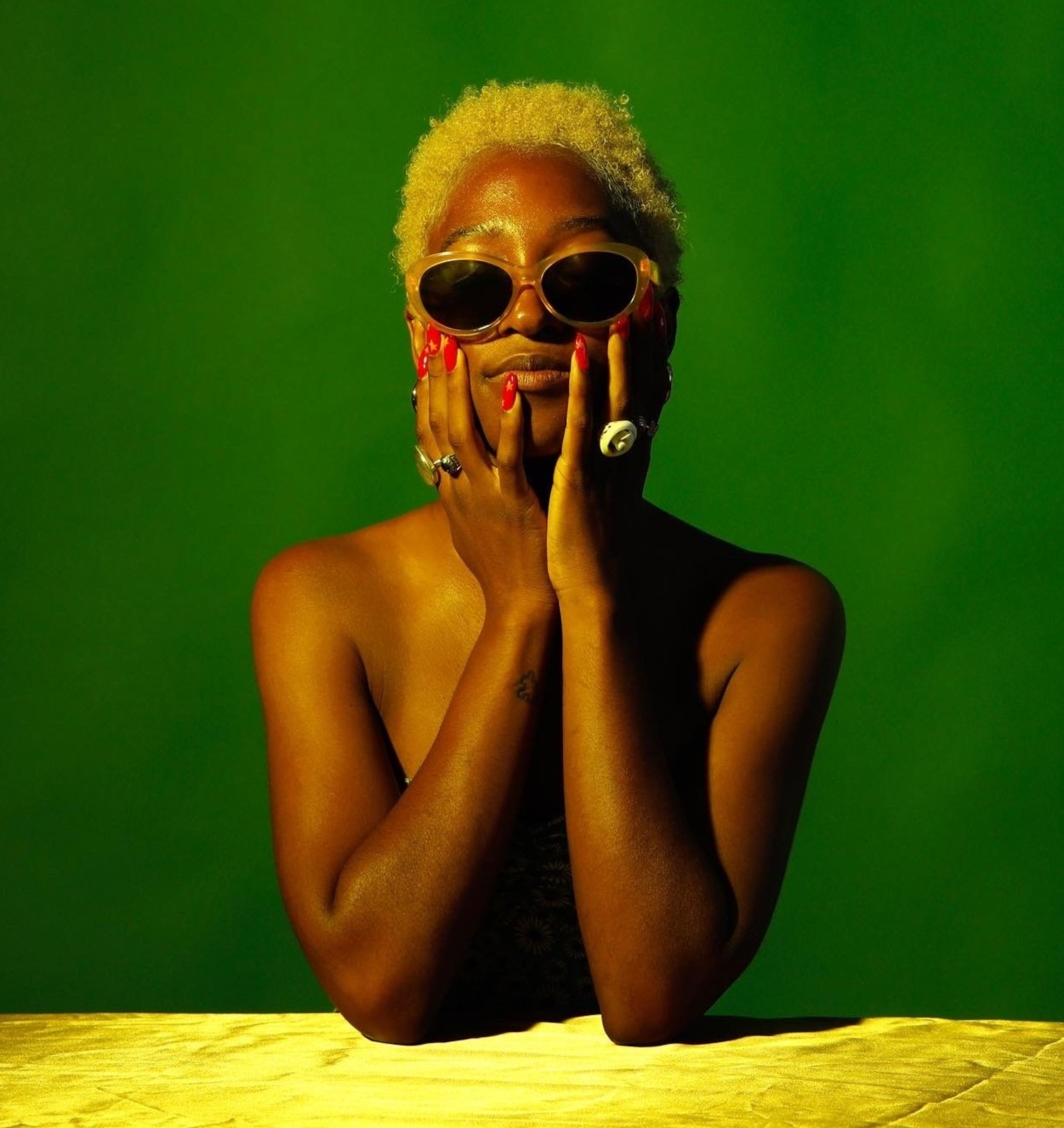 Person with short blonde hair, wearing sunglasses, covering mouth with hands, against a green background.