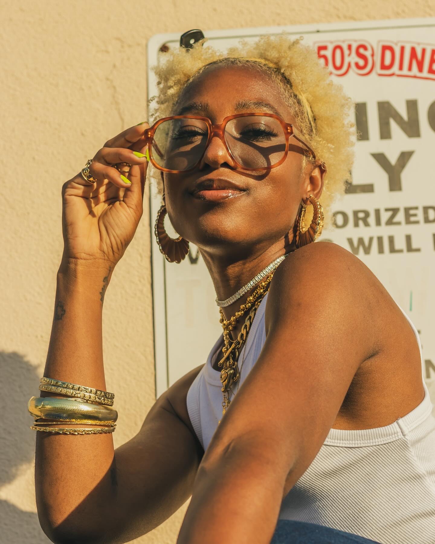 Woman with short blonde hair, glasses, and jewelry, smiling and adjusting her glasses outdoors.