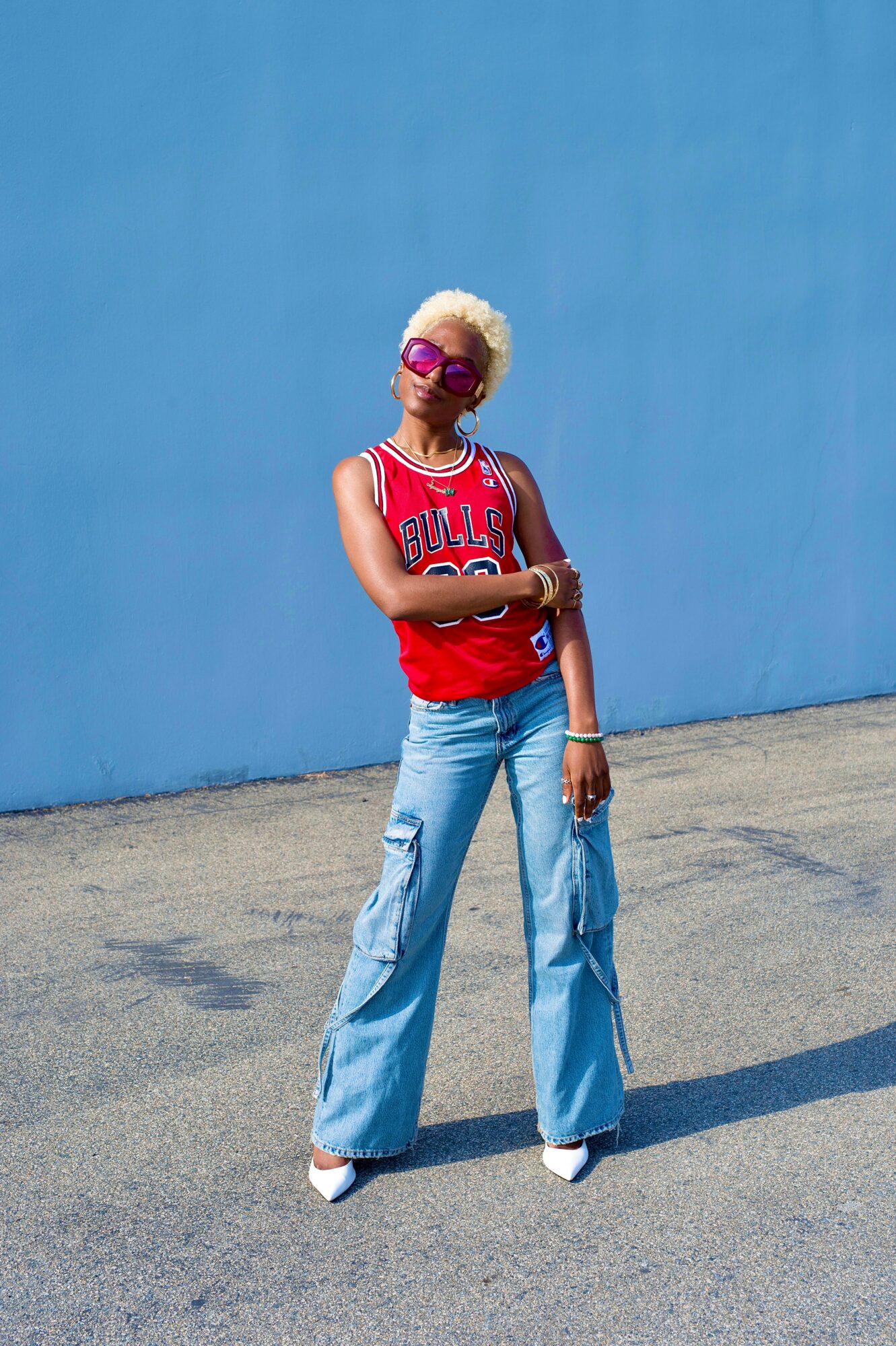Person with short blonde hair wearing sunglasses, a red sports jersey, and wide-leg jeans standing outdoors against a blue wall.
