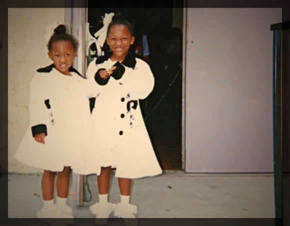 Two young girls in white coats with black accents standing outside a building, smiling, with a door behind them.