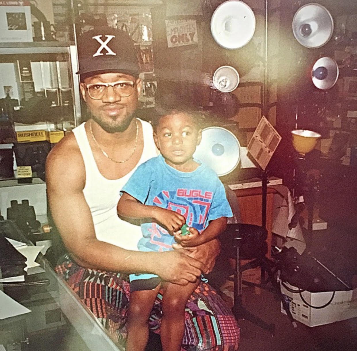 Man and child sitting together indoors, man wearing glasses, cap, tank top, and colorful shorts, child in blue T-shirt, background with speakers.