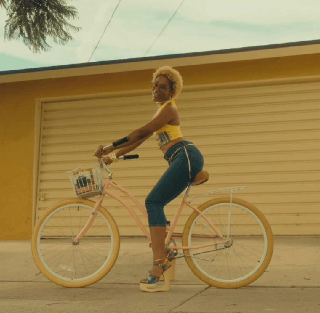 Woman with short blonde hair in yellow top and blue jeans on a pink bicycle in front of a beige garage door.