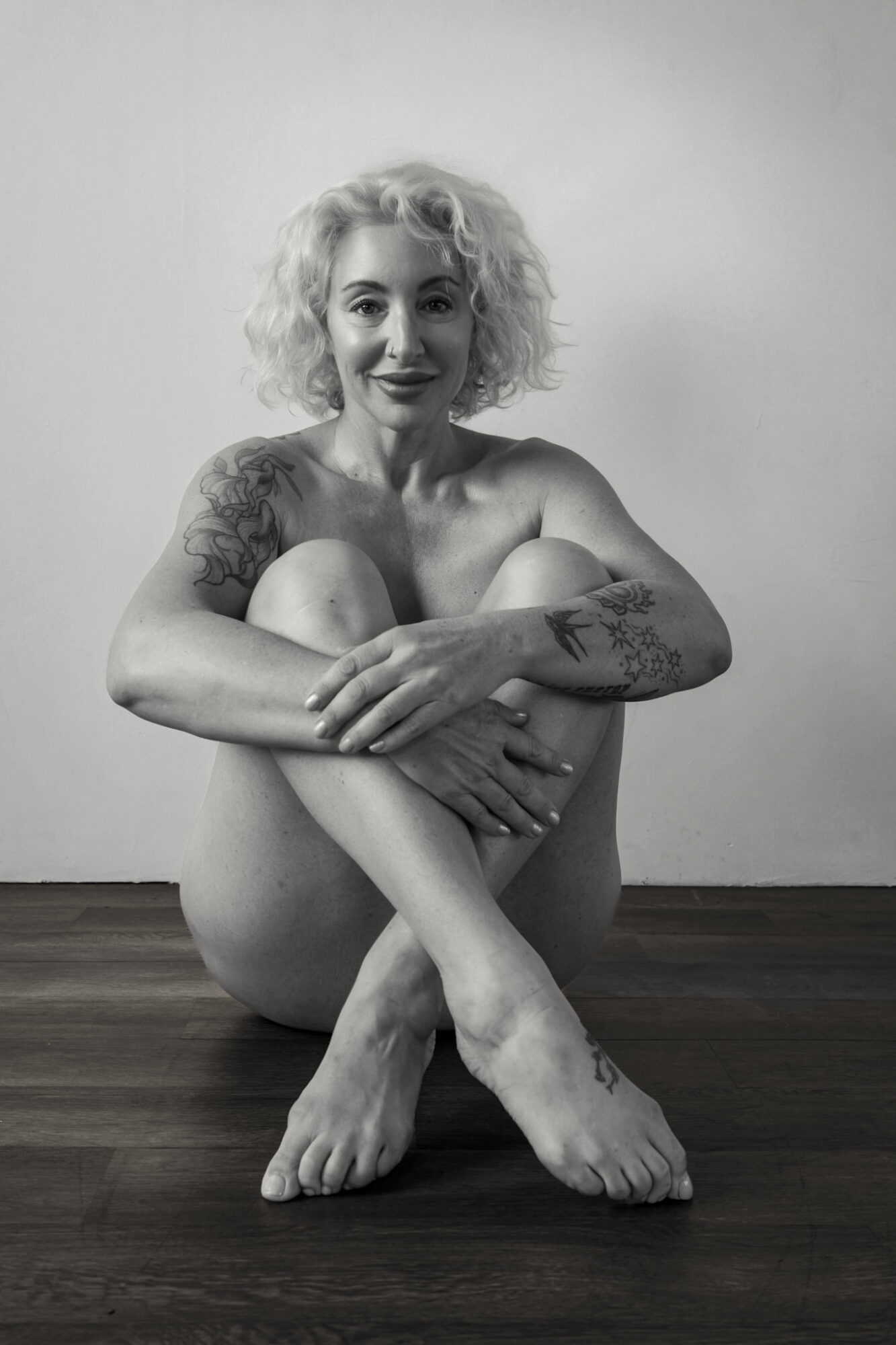 A woman with curly hair sitting on the floor, smiling, with tattoos on her arms, against a plain background.