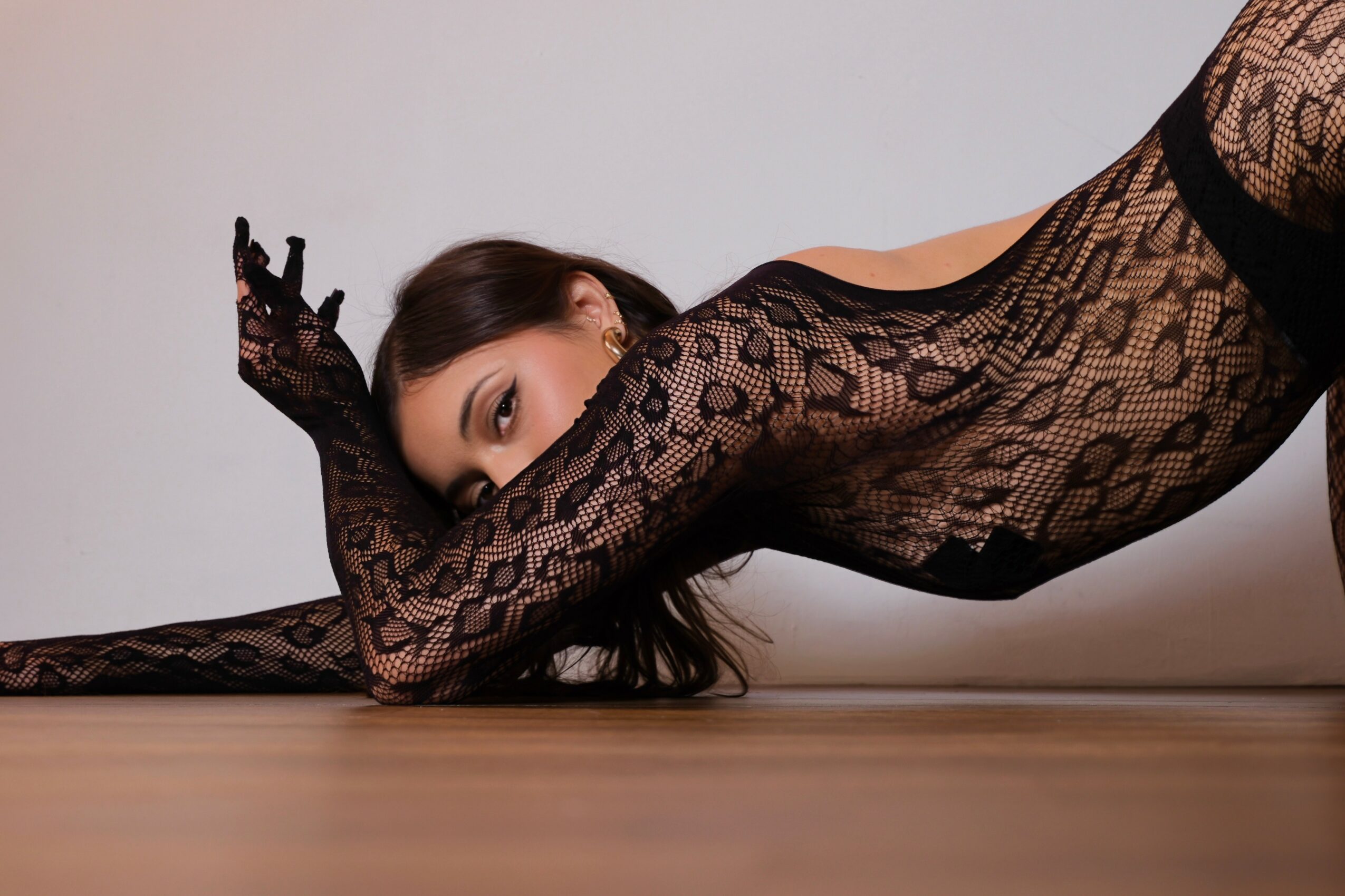 Woman in black lace bodysuit lying on floor, propped on her elbows, looking at camera, with plain background