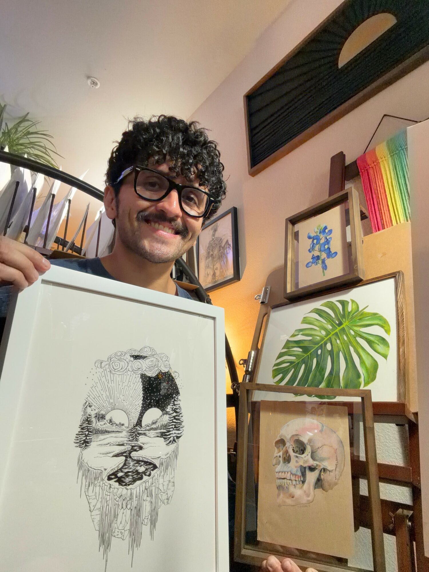 Man with curly hair and glasses smiling, holding framed artwork of a face with a skull, in an art studio with paintings and plants.