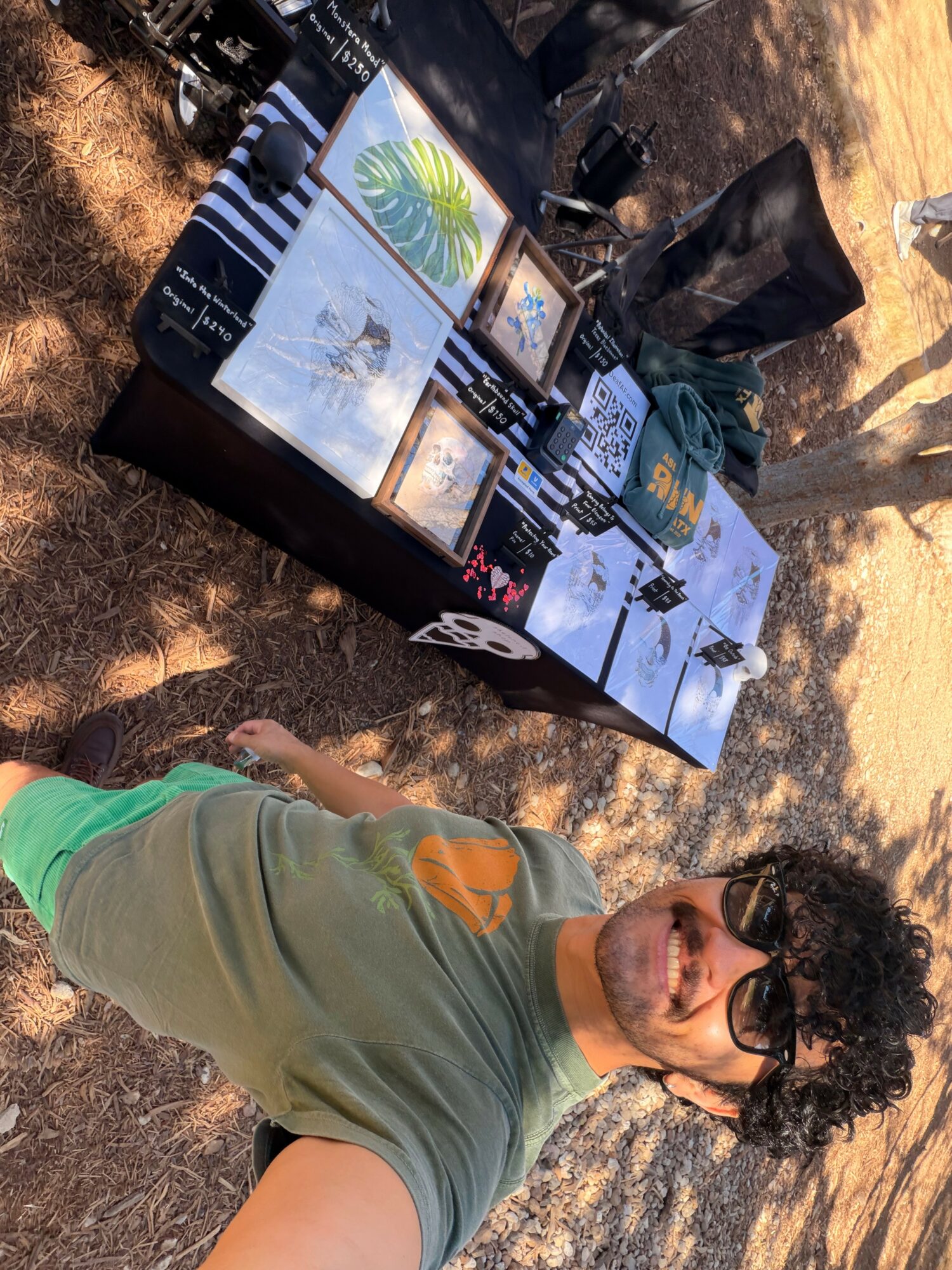 Young man with curly hair and sunglasses smiling outdoors next to a table with artwork and crafts.
