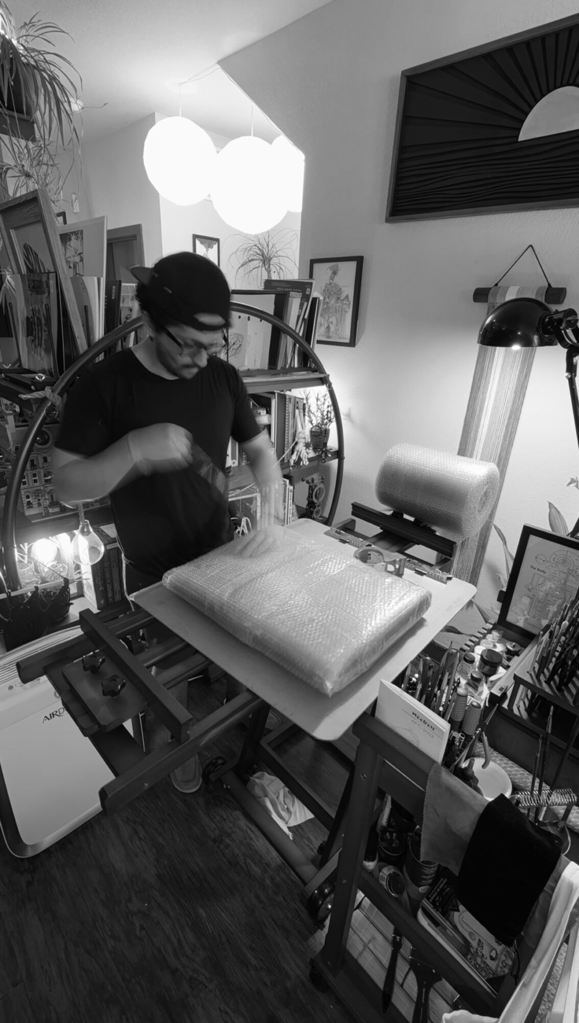 Person wearing glasses and a cap working on a padded surface in a cluttered room with framed pictures and artwork.