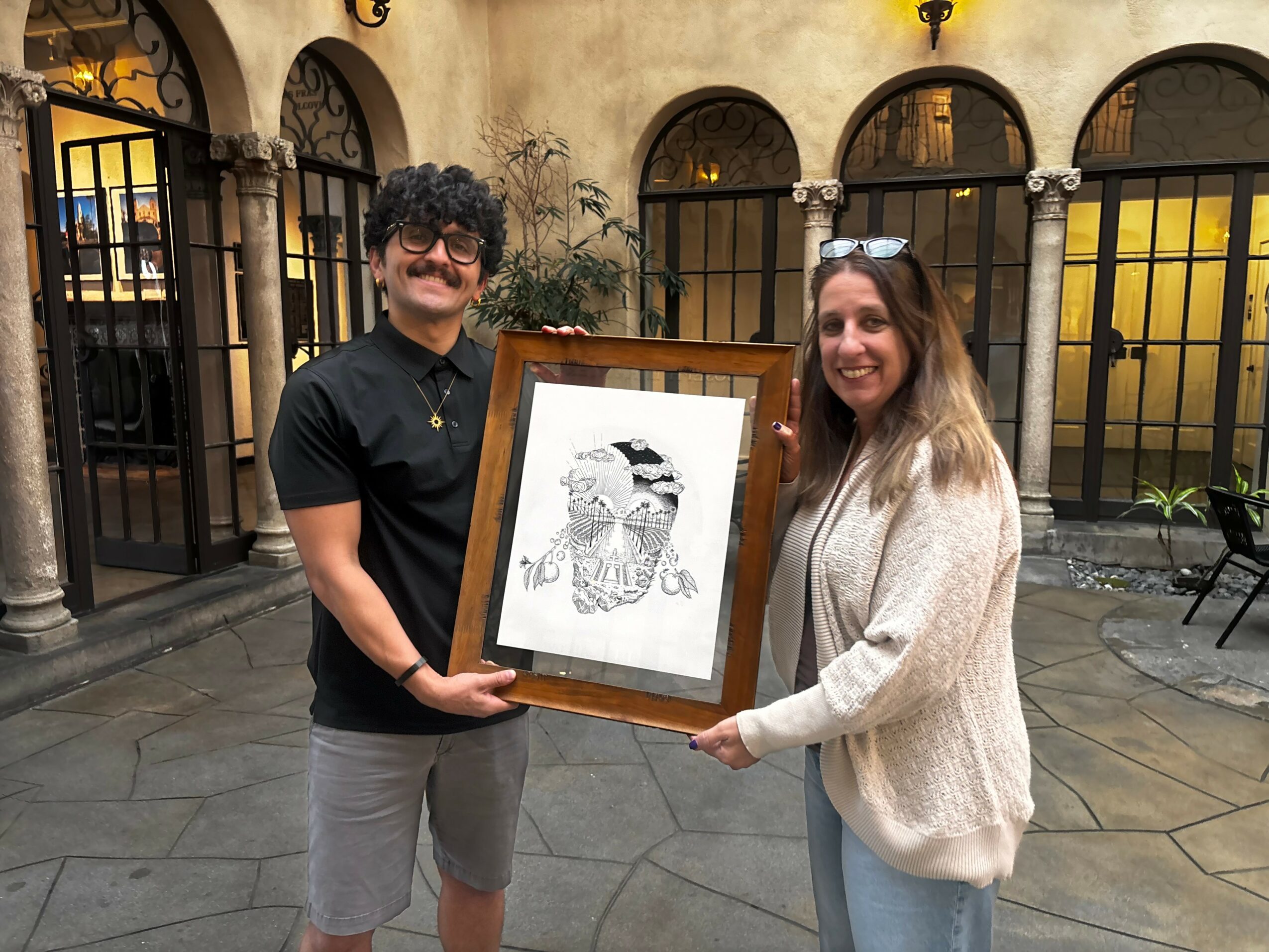 Two people holding a framed black and white drawing, standing outdoors in a courtyard with arches and iron gates.