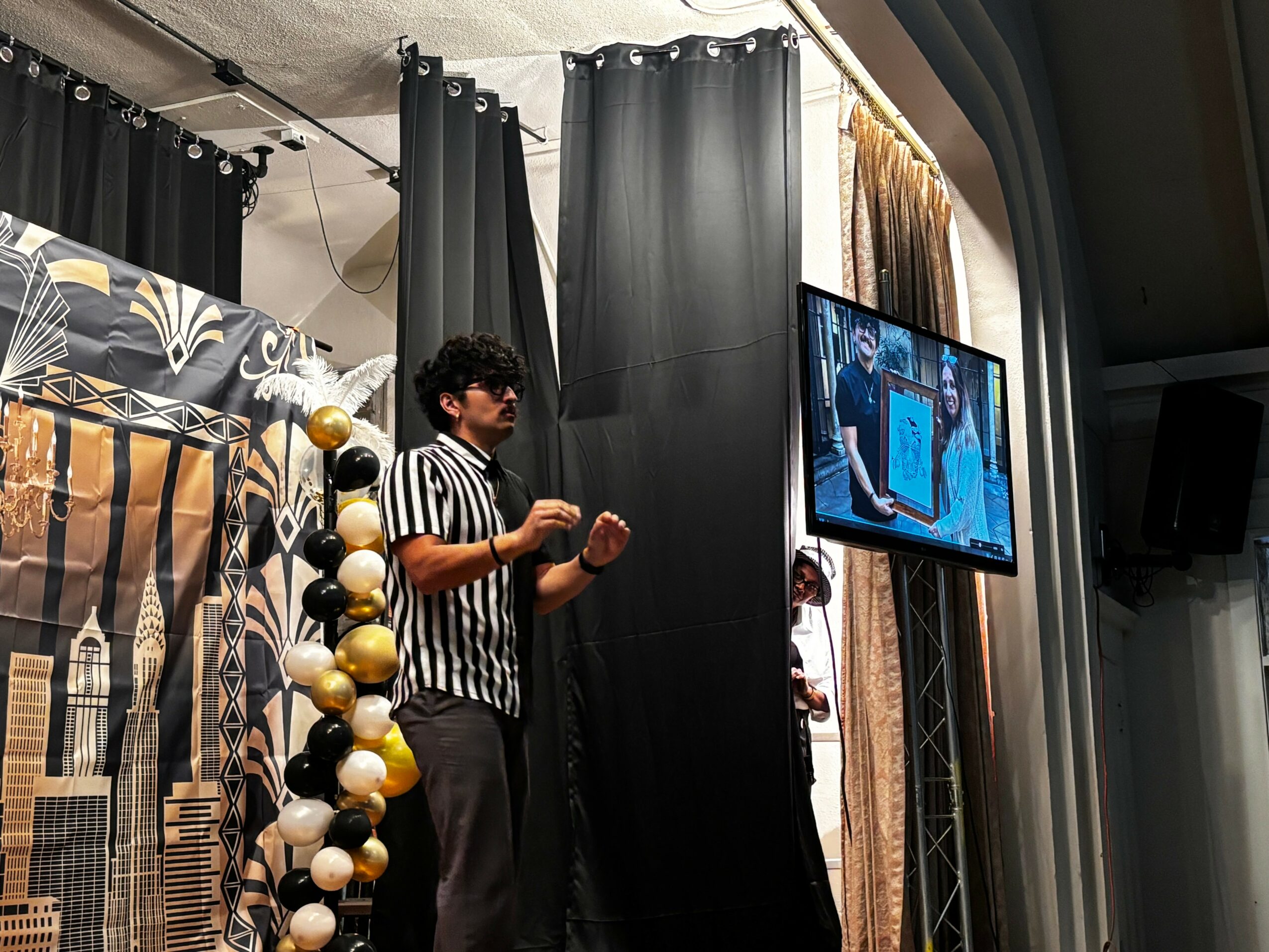 Person standing on stage with black curtains, decorated with gold and white balloons, next to a large screen displaying a presentation.