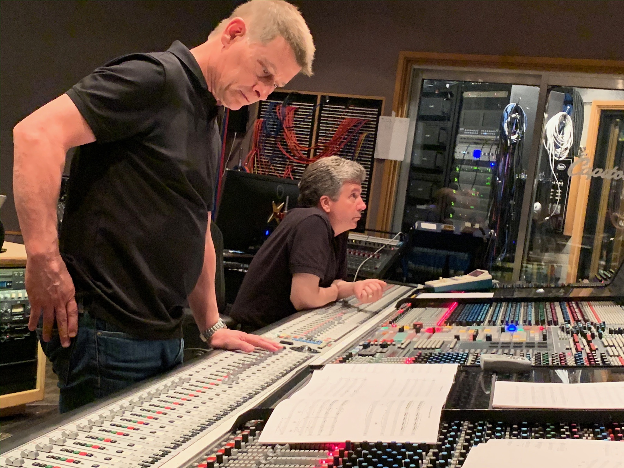 Two men working at a large audio mixing console in a recording studio, one standing and one seated, with equipment and soundproofing.