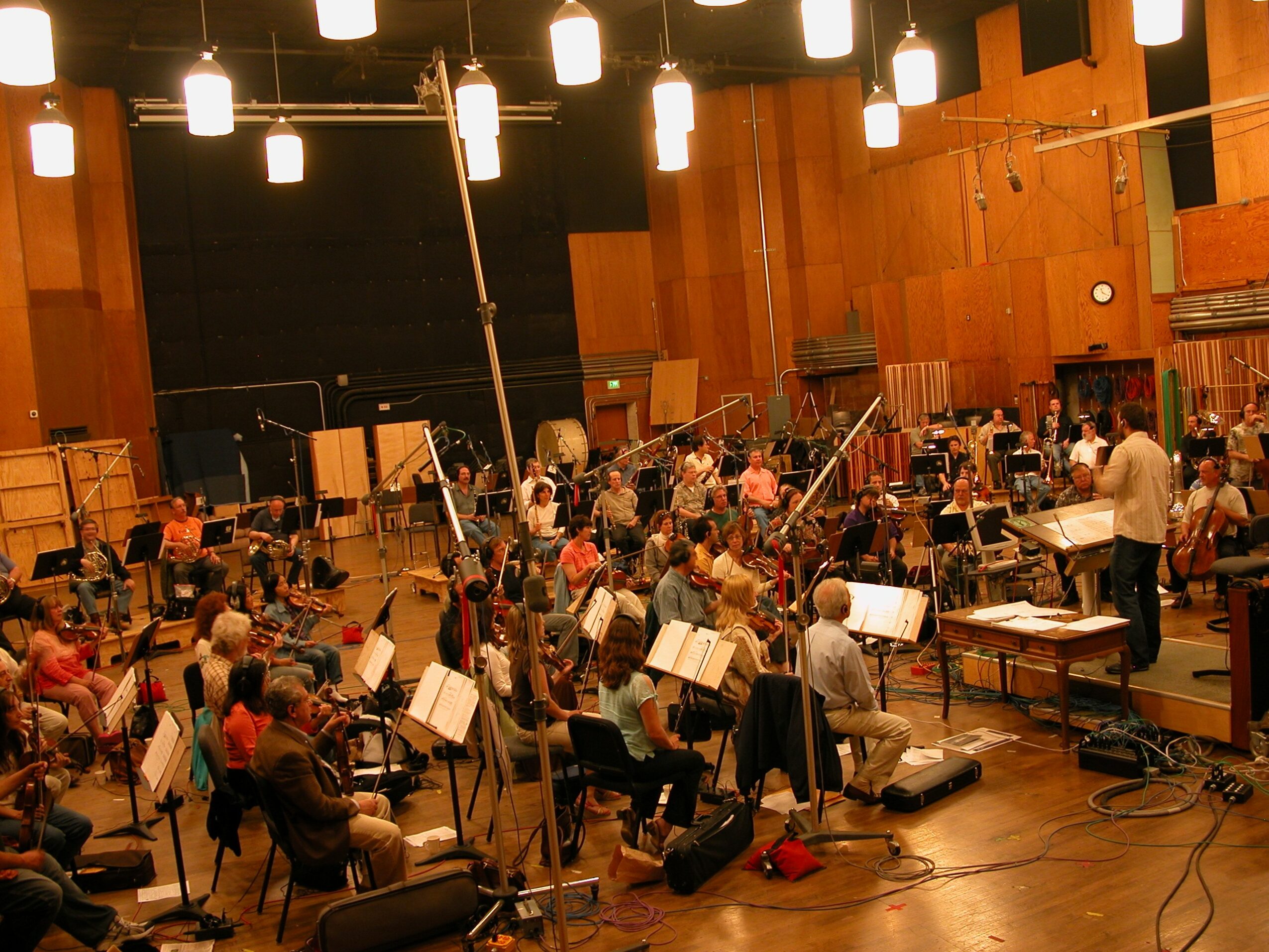 Orchestra rehearsal with musicians, conductor, and music stands in a large wooden auditorium.