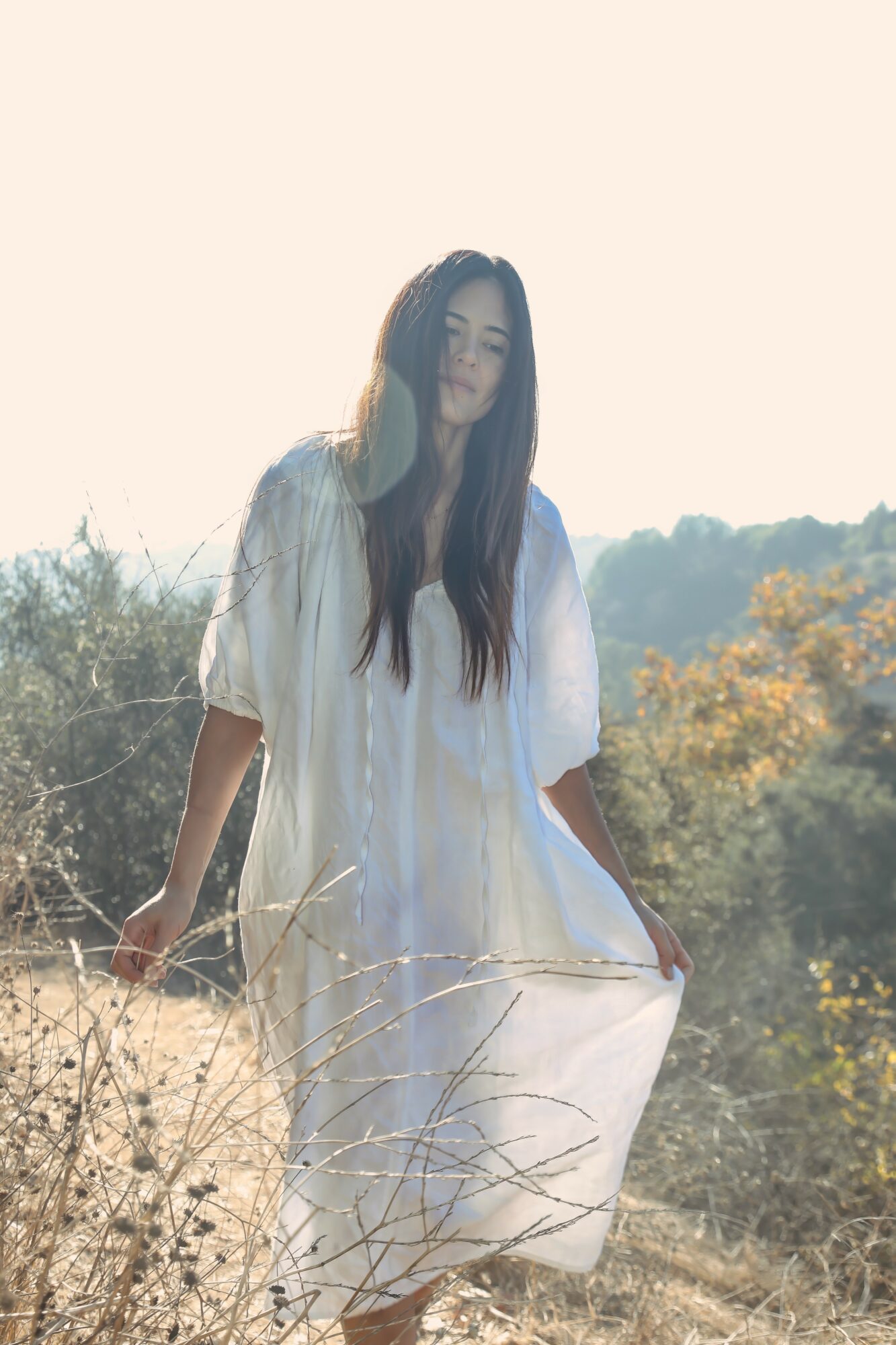 Woman in white dress standing outdoors with trees and sky in background, holding the dress with one hand.