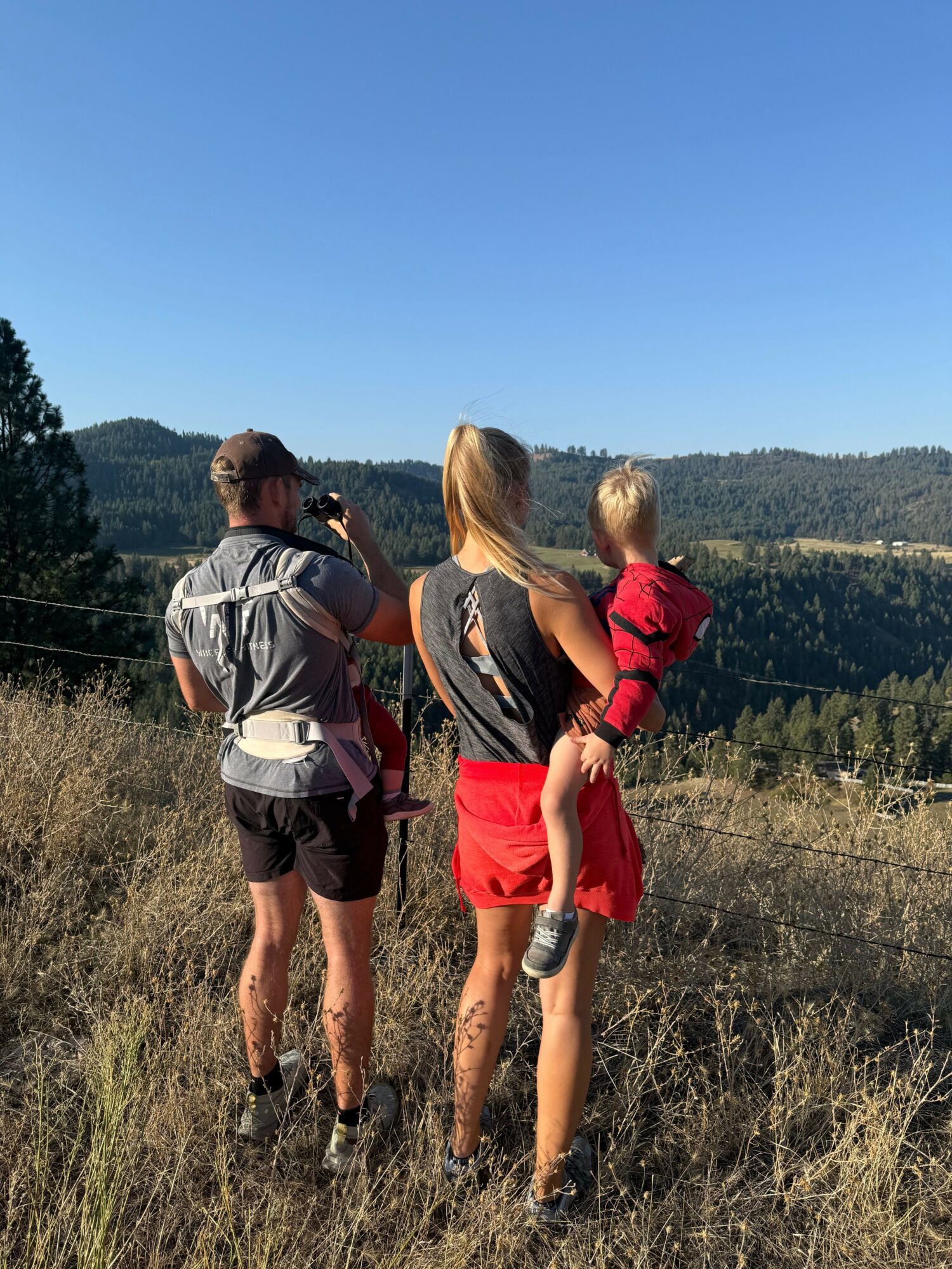Two people walking on a trail in a natural landscape with trees and hills, under a clear blue sky.