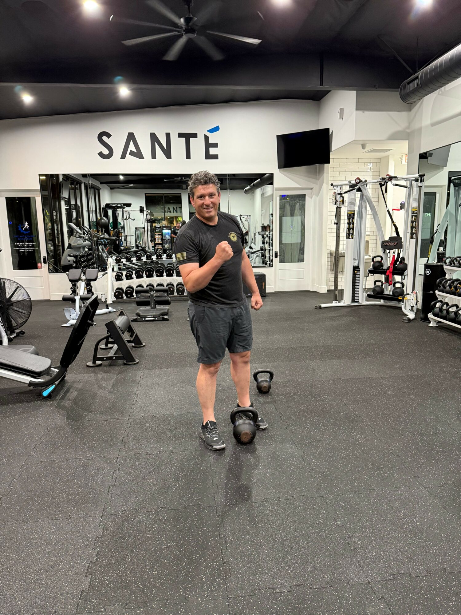 A man stands in a gym with weightlifting equipment, smiling, wearing a black t-shirt and shorts, with a gym logo in the background.