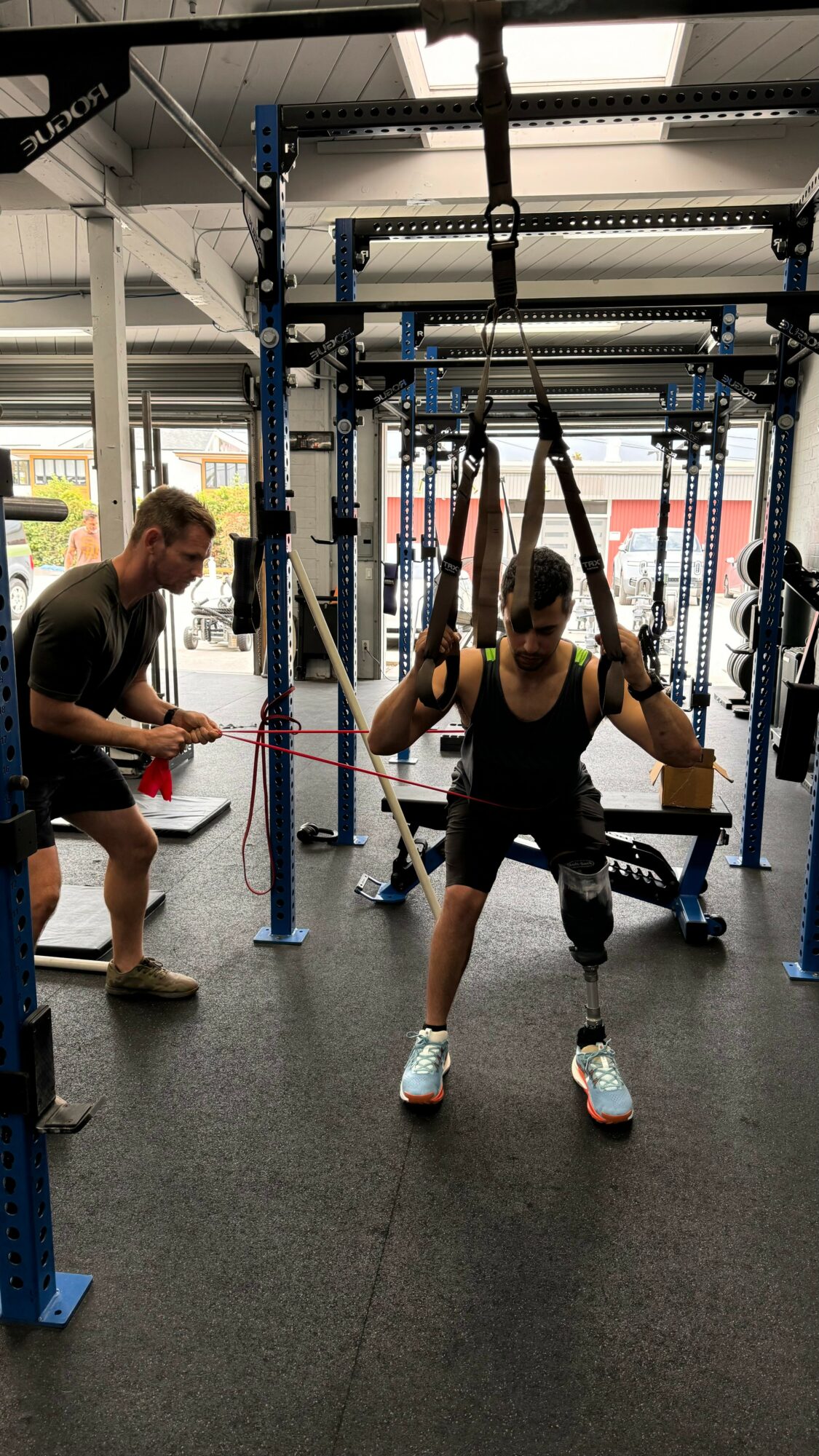 Person using suspension straps for exercise in gym with others nearby, industrial-style interior, black flooring, and metal equipment.