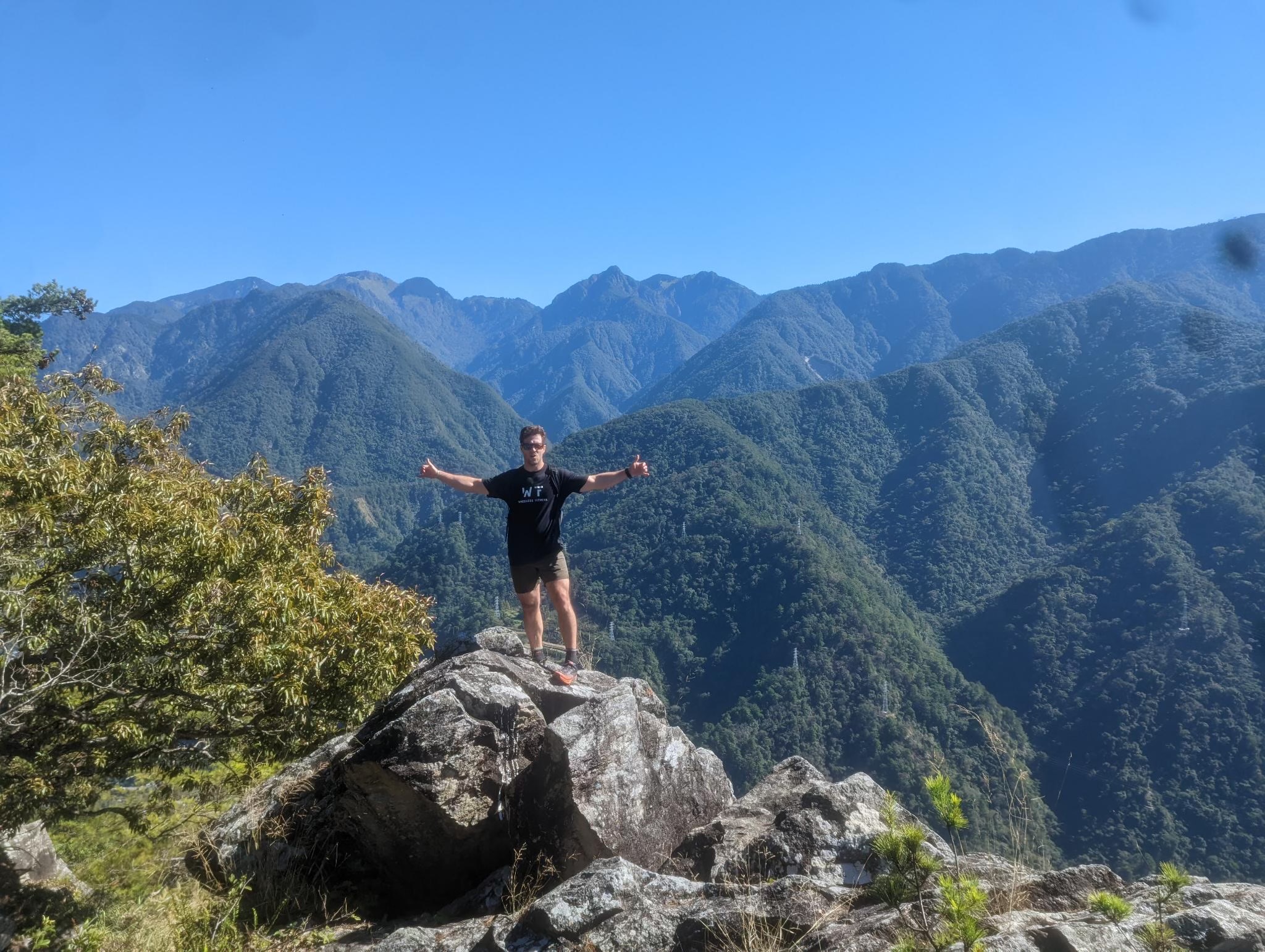 Person standing on large rock with arms outstretched, mountains in background, clear blue sky, trees in foreground.