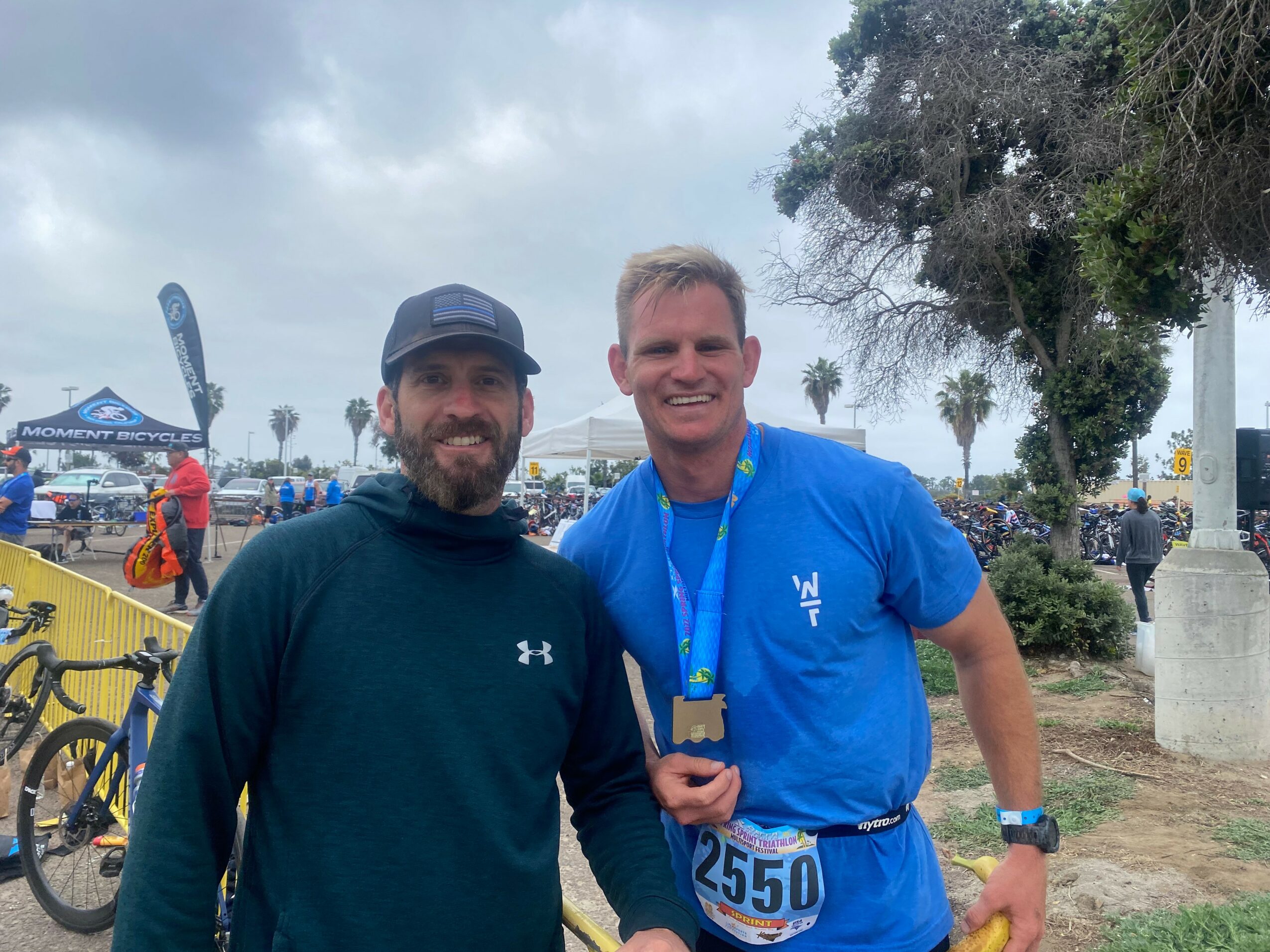 Two men smiling outdoors, one wearing a dark hoodie and the other in a blue shirt with a medal, with trees and people in the background.