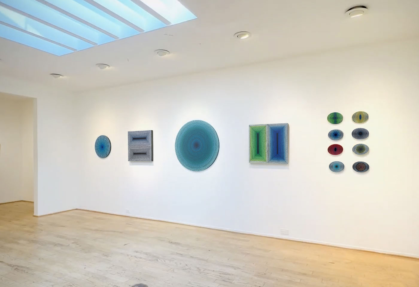 Gallery wall with colorful circular and rectangular abstract art pieces on a white wall, wooden floor, and ceiling lights.