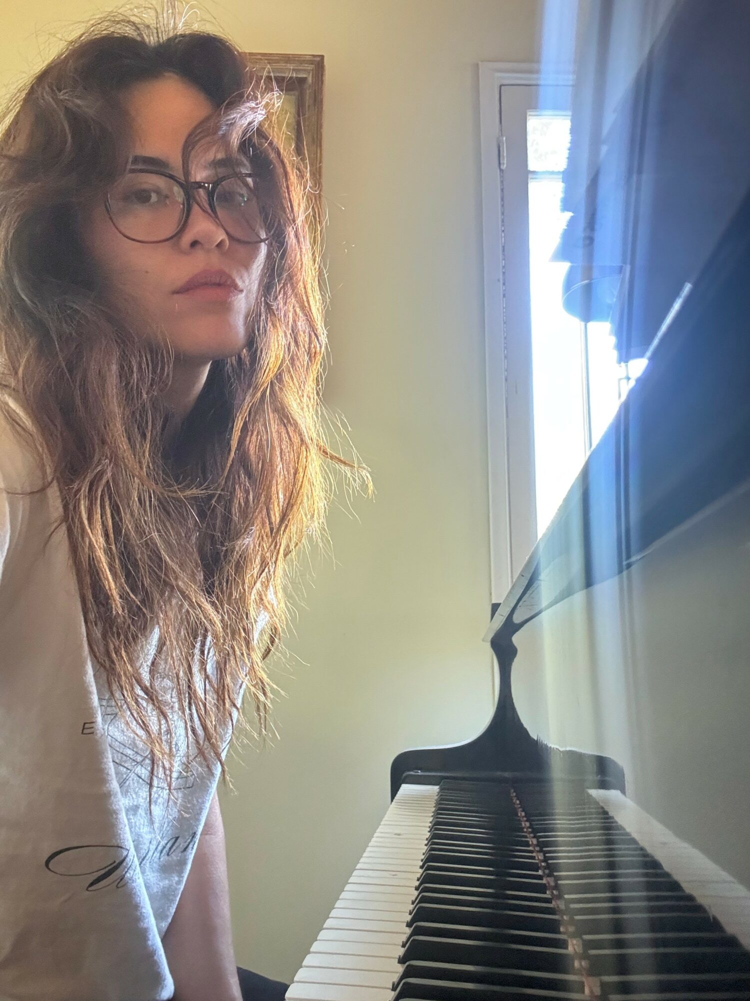 Young woman with glasses and wavy hair sitting at a piano, looking at the camera, with a window in the background.