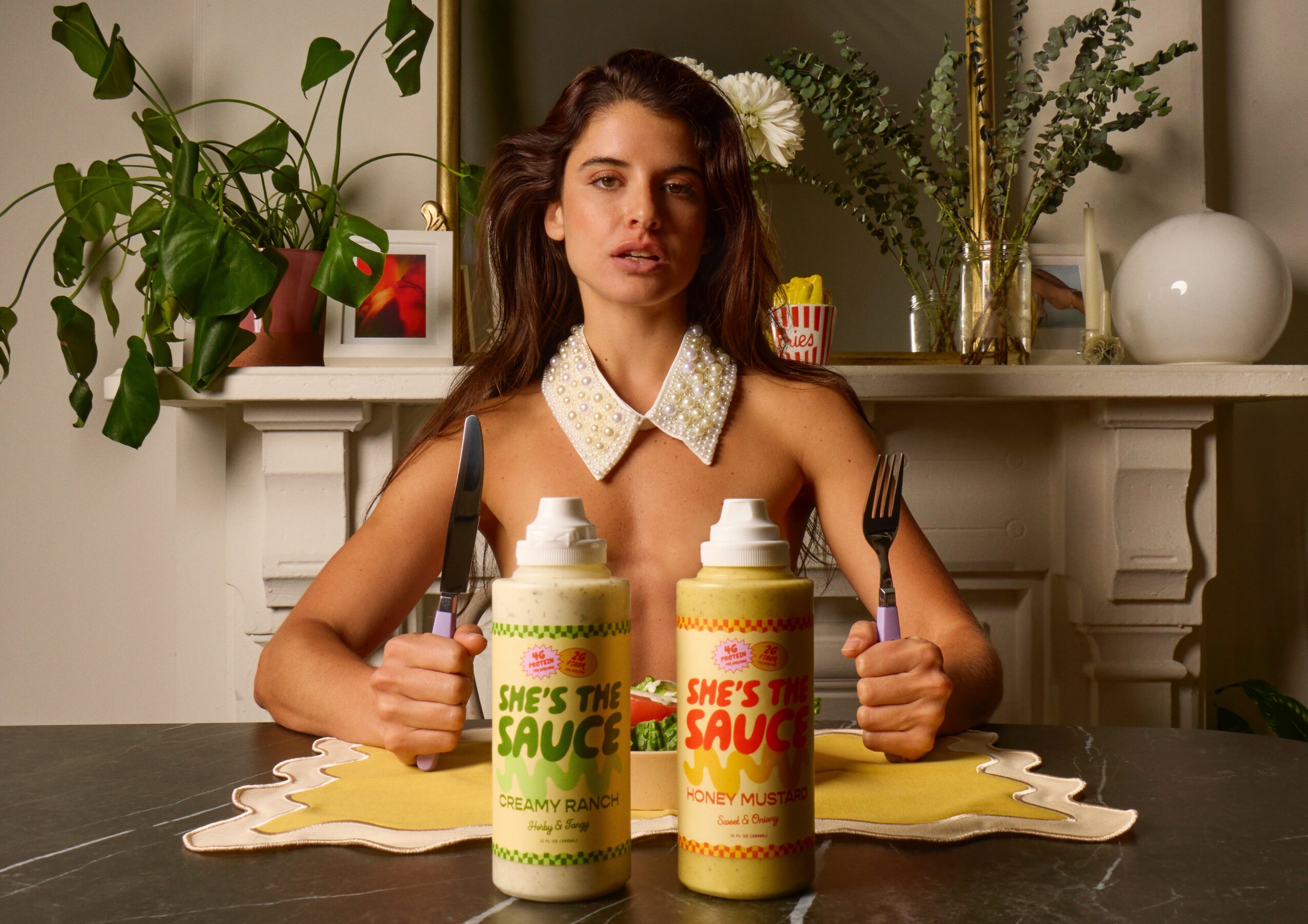 Young woman with wavy hair sitting at a table with two bottles of sauce, holding a fork and knife, in a decorated room.