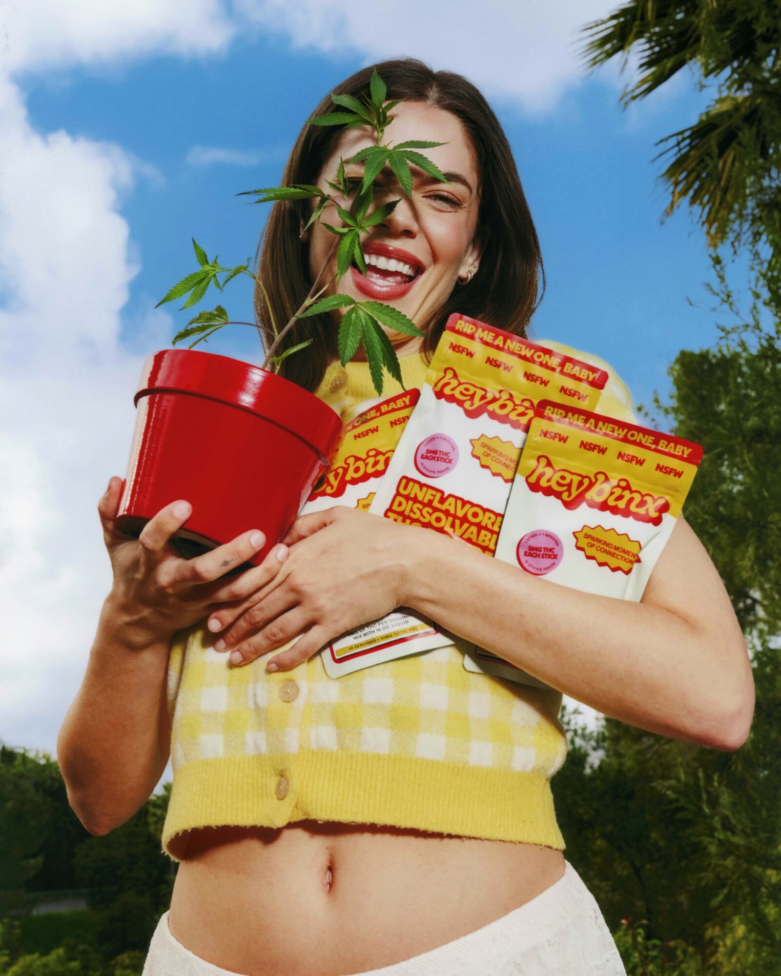 Smiling woman holding a potted plant and snack packages outdoors with trees and blue sky.