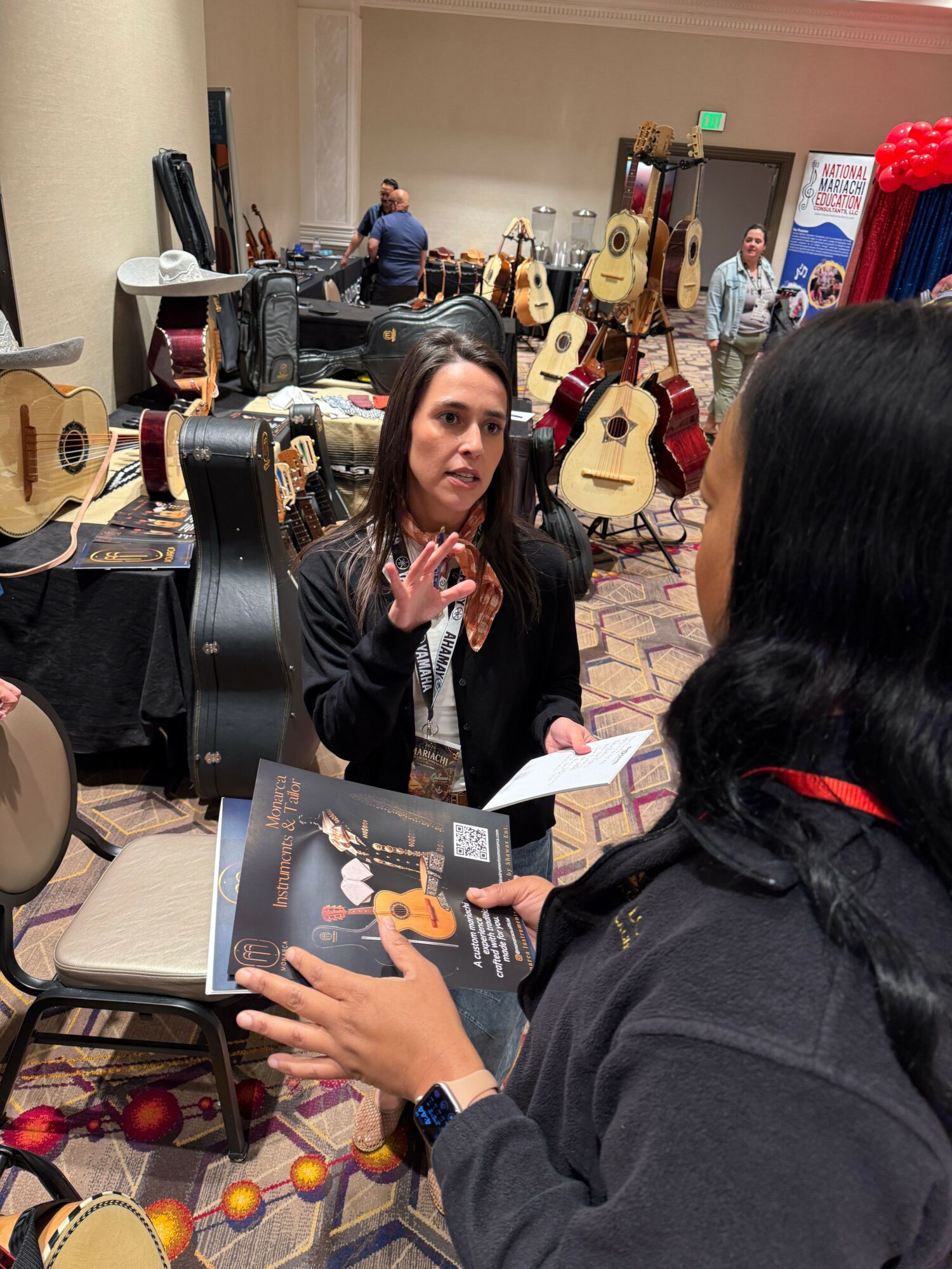Woman sitting at table with guitars displayed behind her, talking to a person holding a music book, in a store or event space.